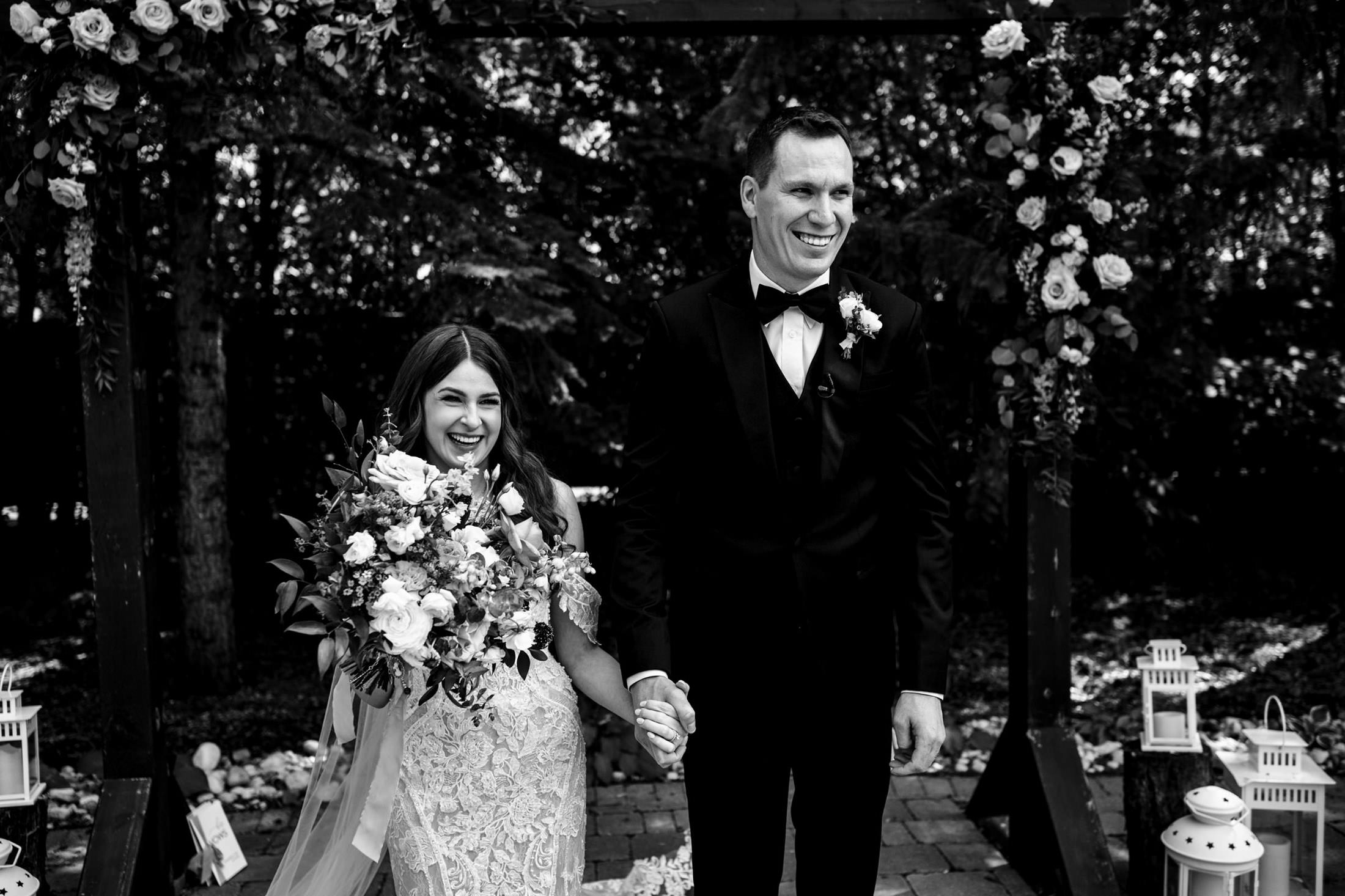 Bride and groom smiling, holding hands under a floral arch at Ashgrove Acres Wedding.
