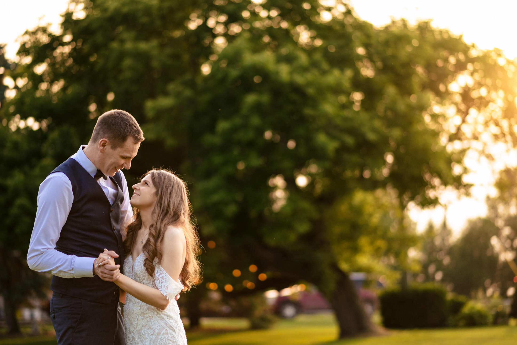 A couple dances at Ashgrove Acres, under trees at sunset, holding hands and smiling.