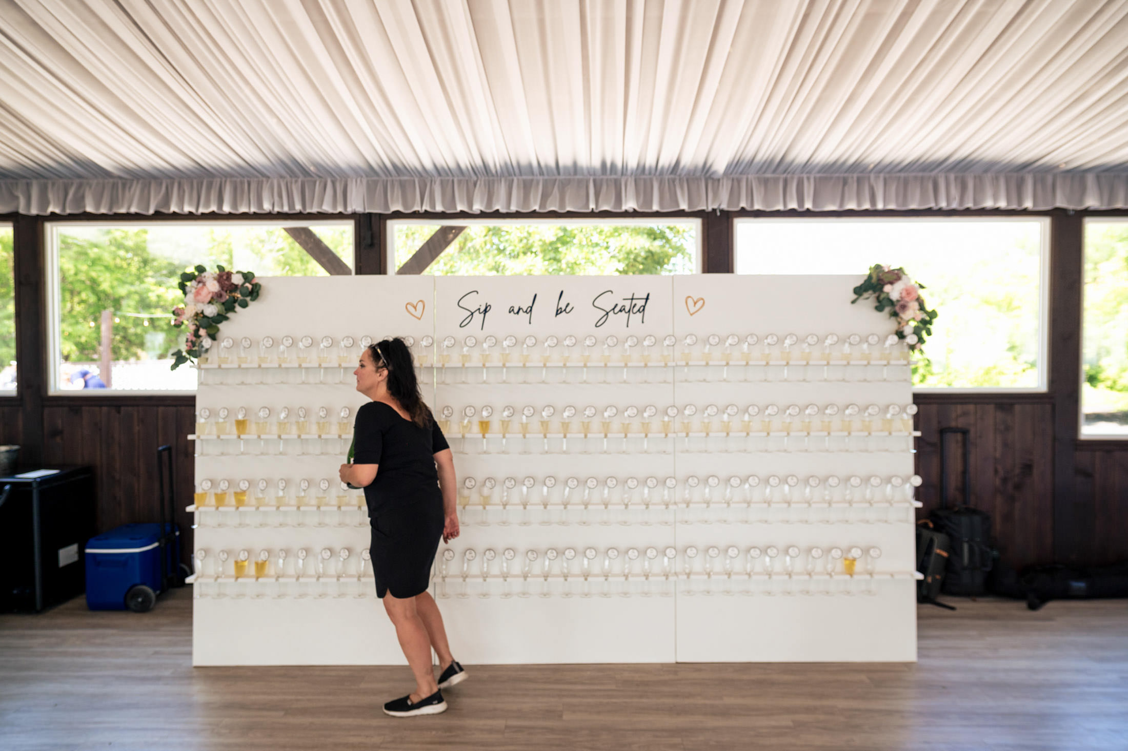 Woman strolling past champagne wall with "Sip and be Seated" sign at Ashgrove Acres Wedding.