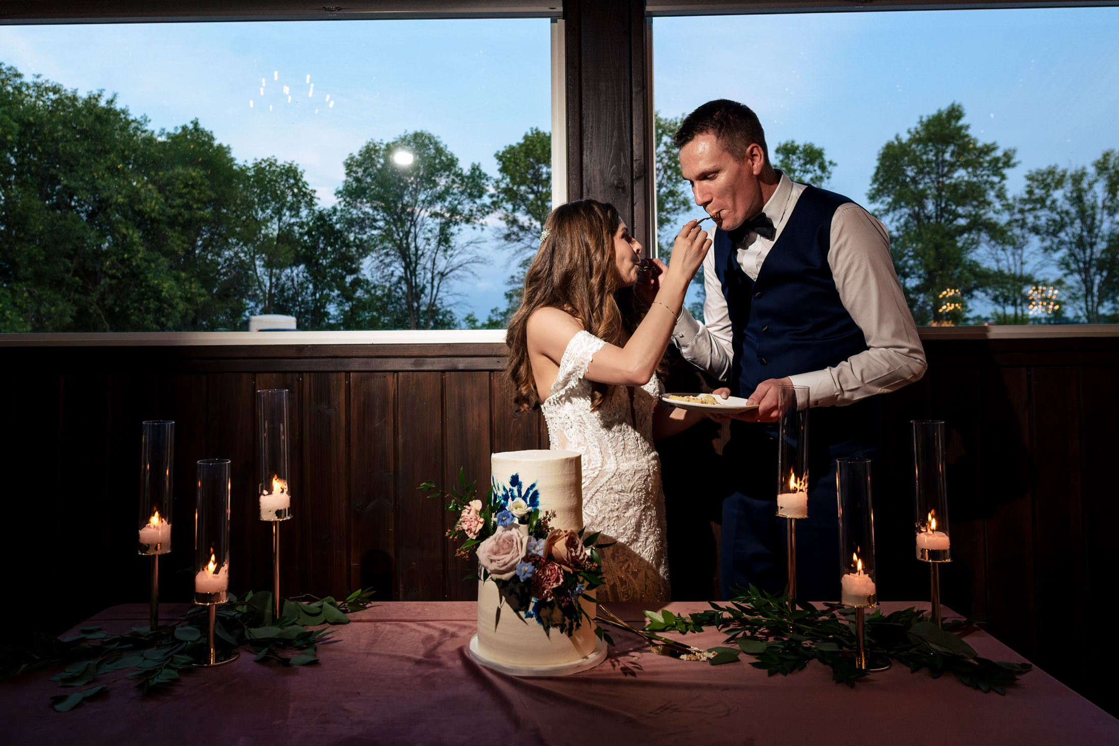 Bride and groom at Ashgrove Acres feeding each other cake by candlelit table.