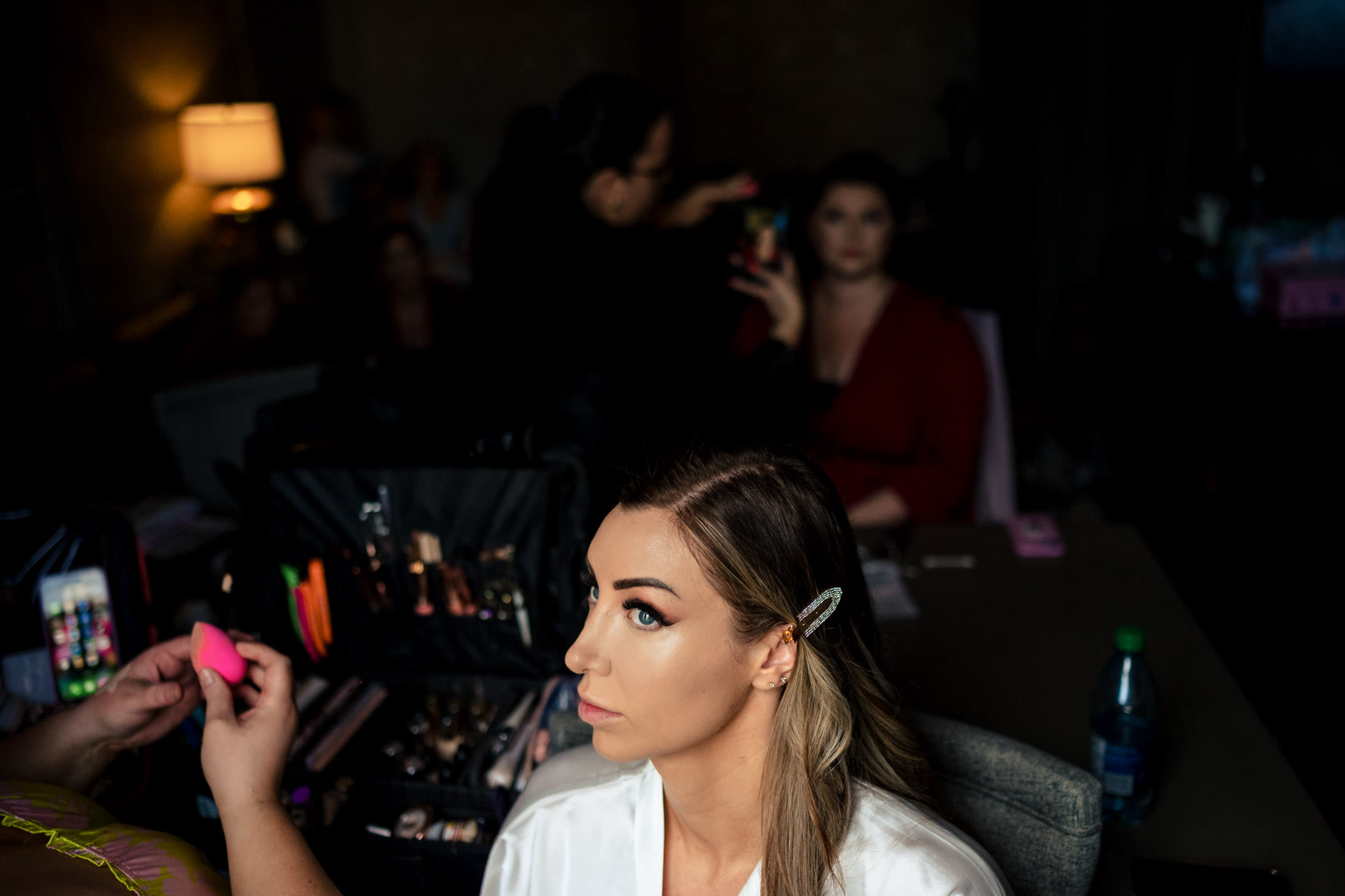 Woman getting makeup for an Italian wedding, seated with others in a dimly lit room.