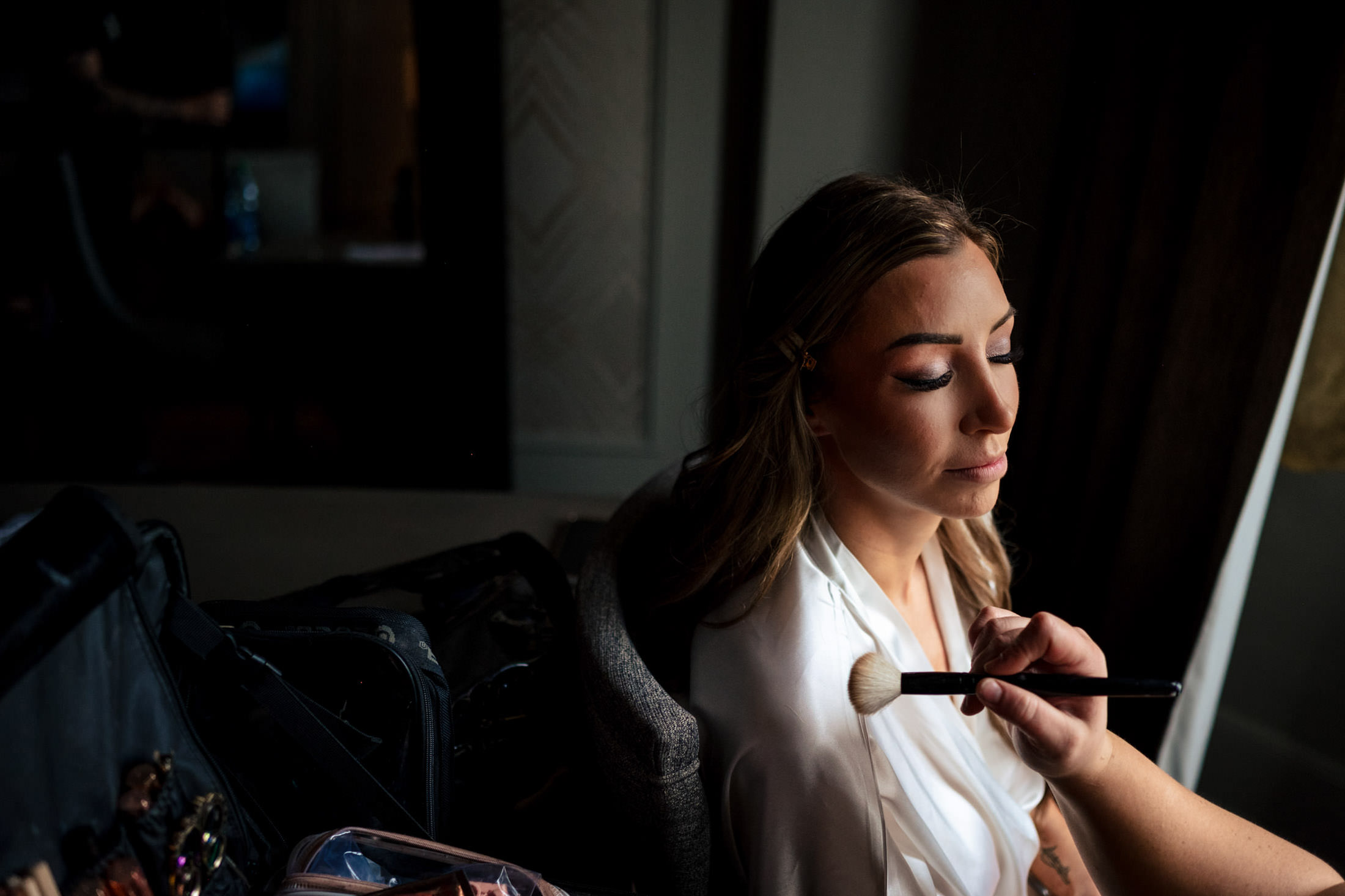 Woman in a white robe getting makeup for an Italian wedding in a dimly lit room.