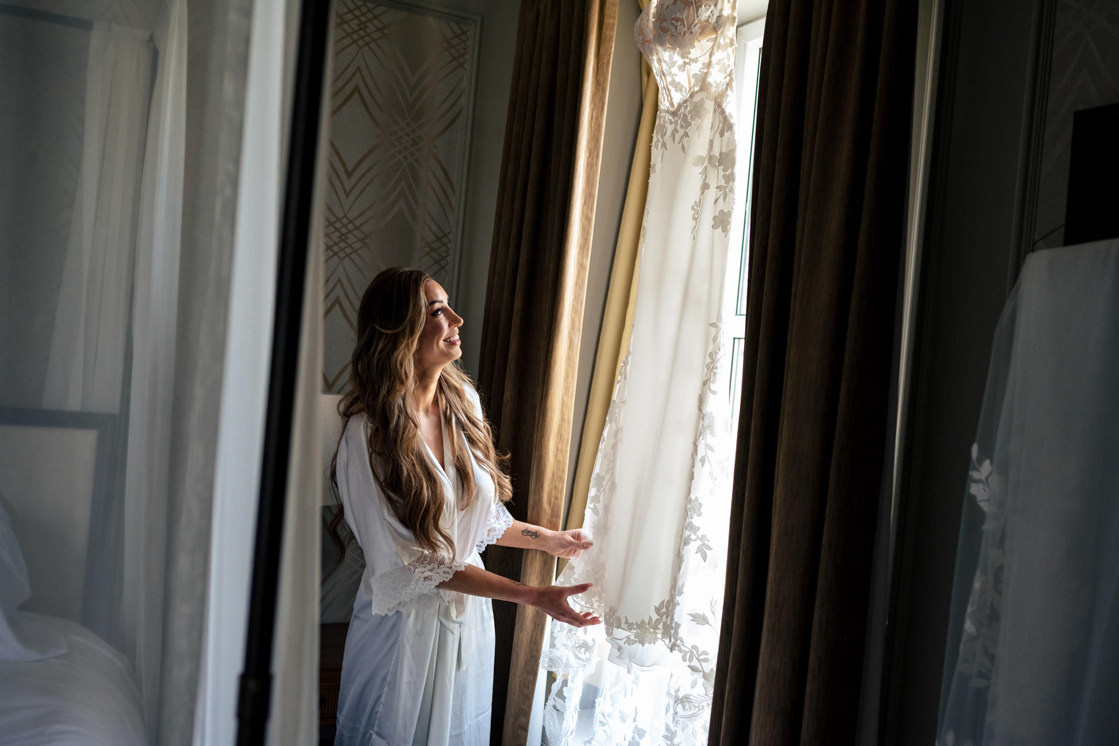 Woman in robe smiles, holding a wedding dress by a window at an Italian wedding.