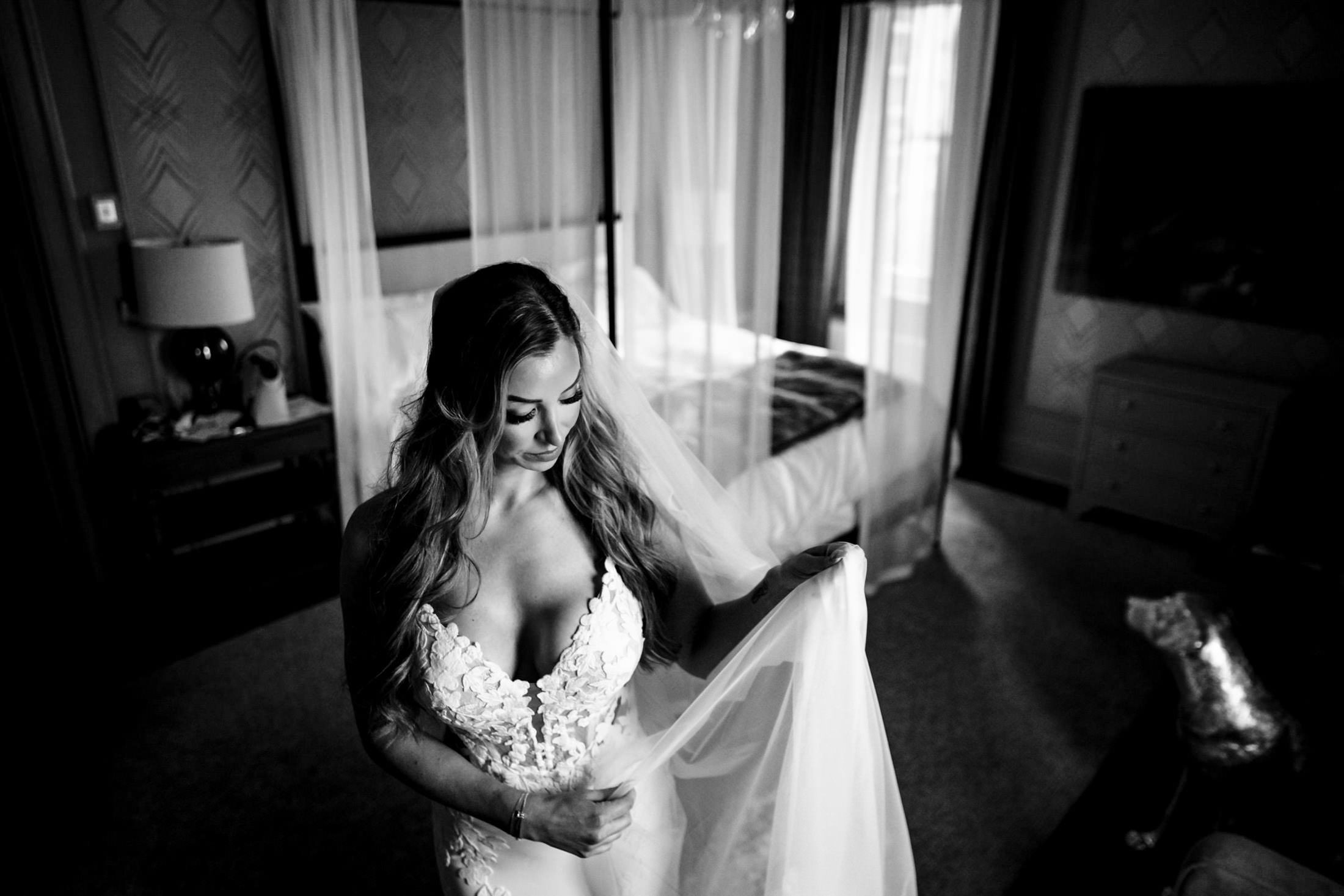 Bride in wedding dress adjusts her veil in an elegant Winnipeg bedroom. Black and white photo.