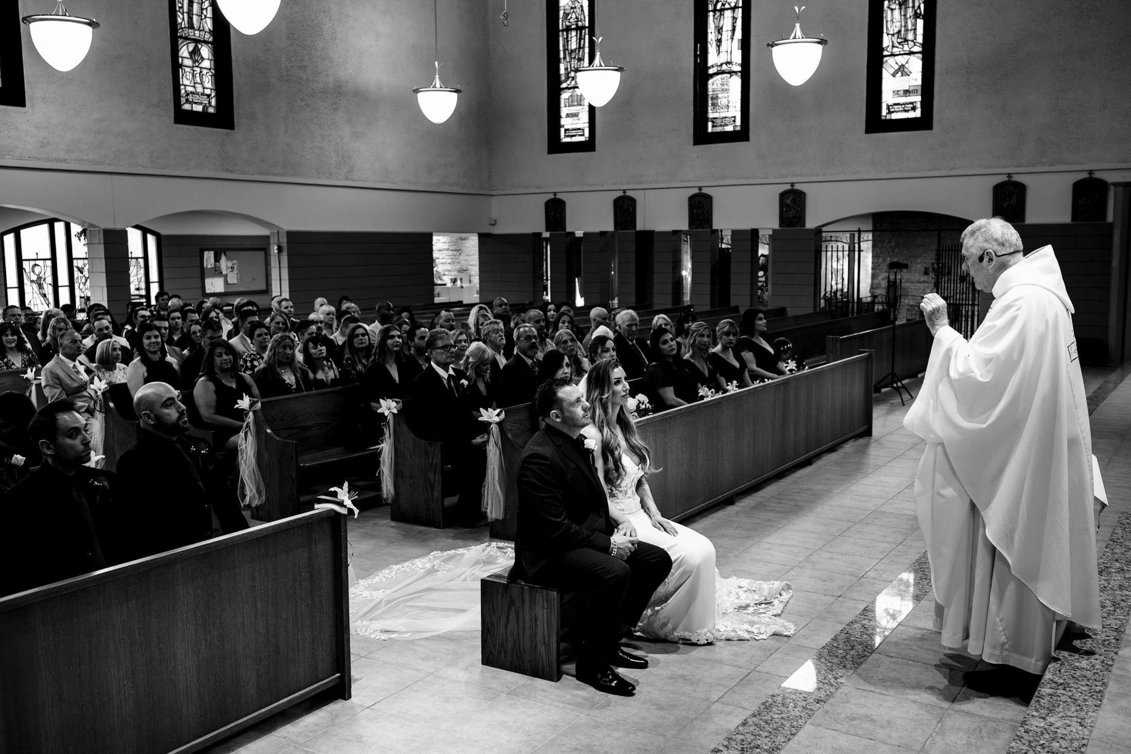 A couple seated at their Italian wedding in Winnipeg, with a priest conducting the ceremony.