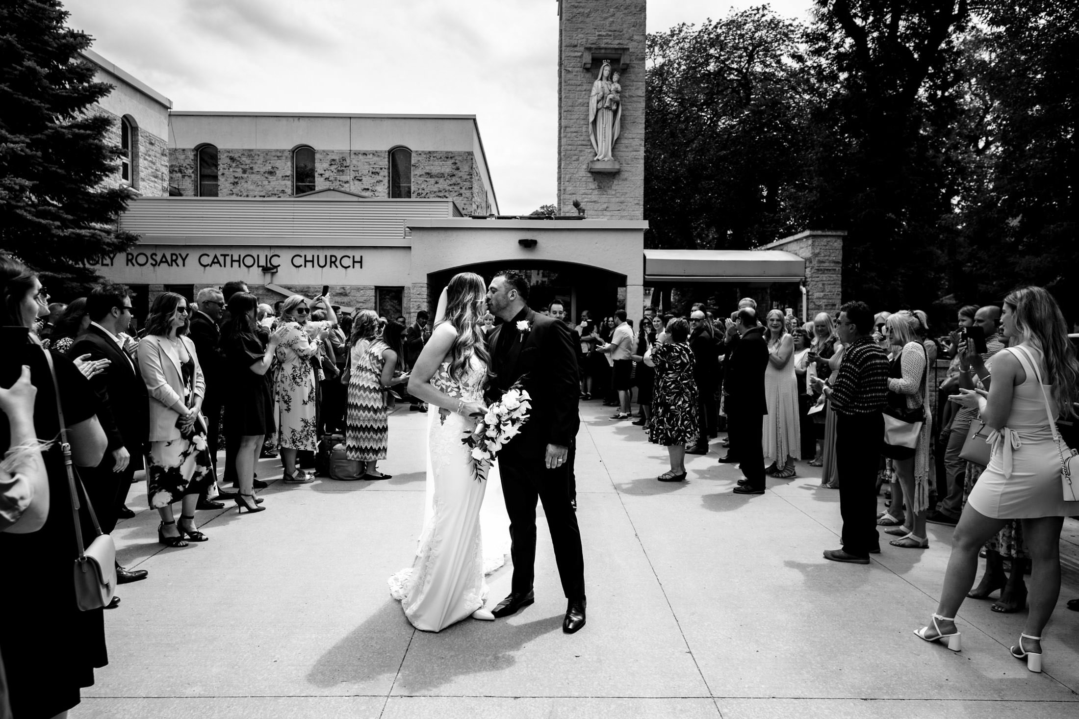 The bride and groom kiss outside a church in Winnipeg as guests celebrate their Italian wedding.