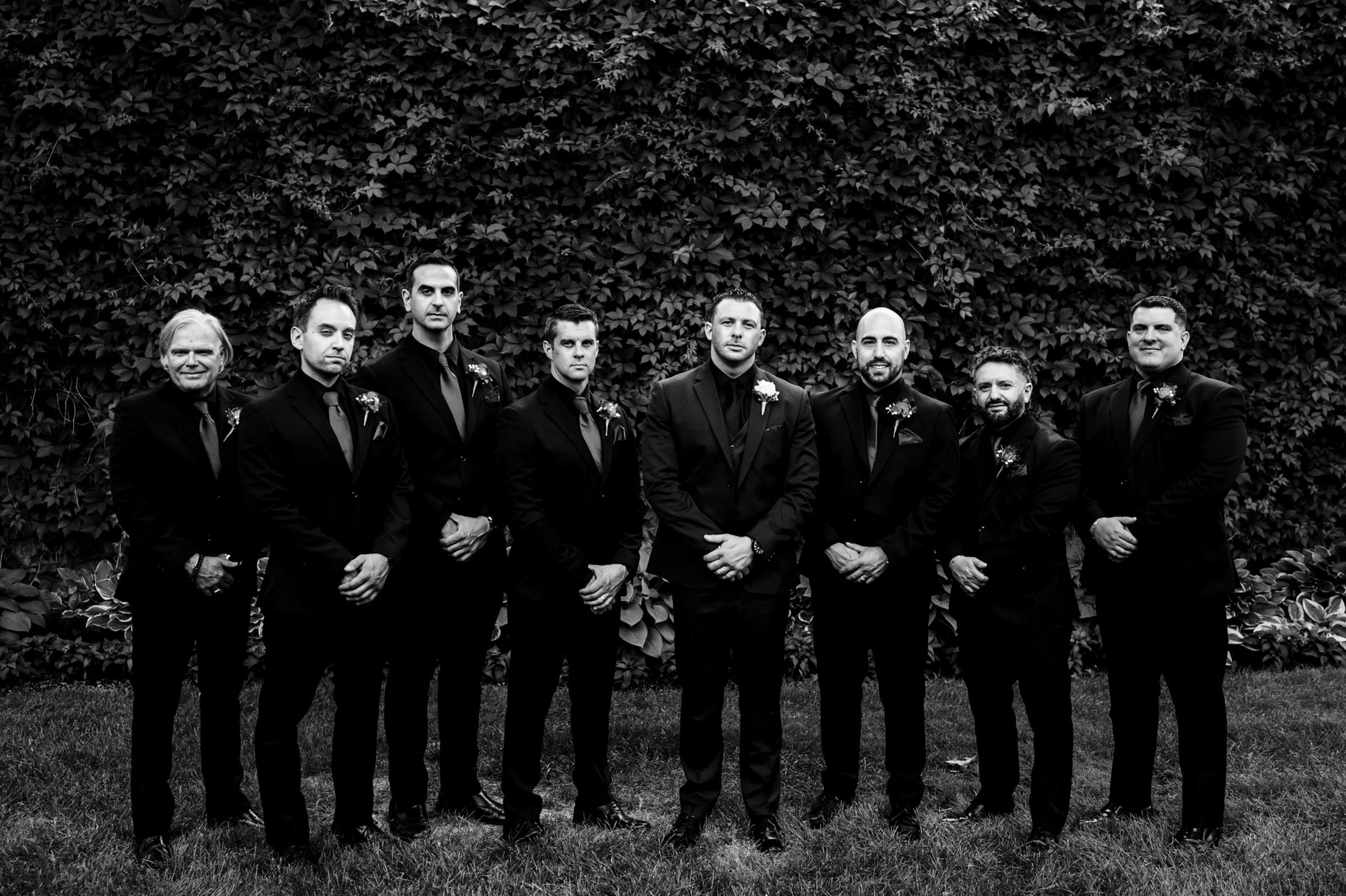 Eight men in suits pose for a group photo at an Italian wedding in front of a leafy wall.