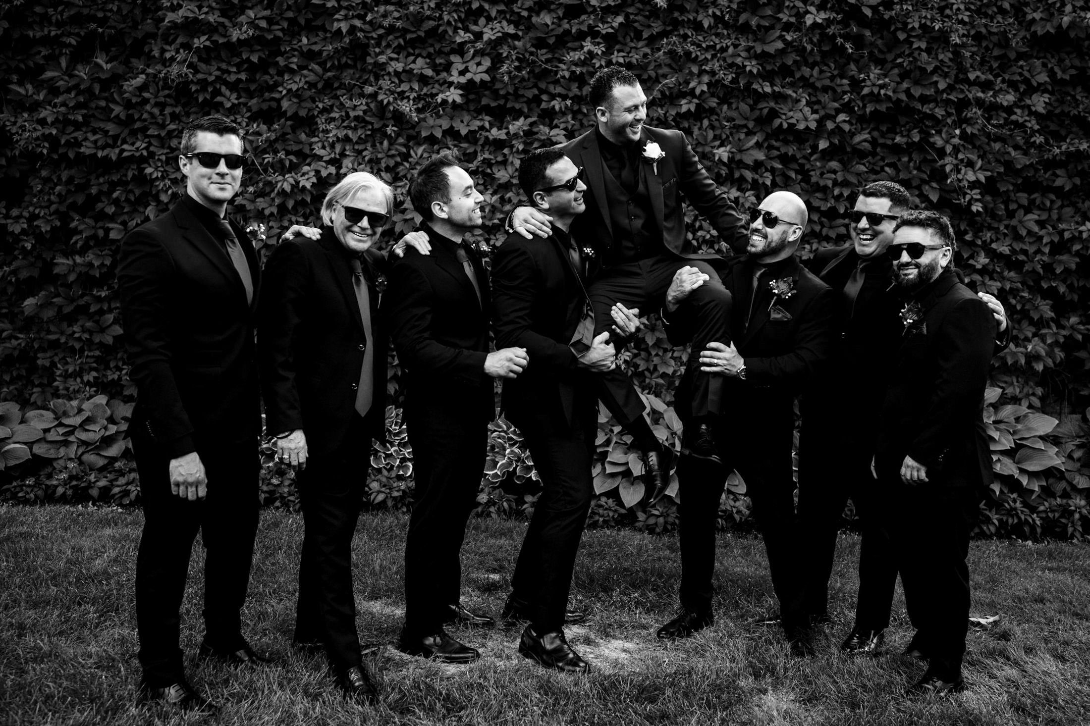 Men in suits lift one man, smiling at an Italian wedding in Winnipeg before a vine-covered wall.