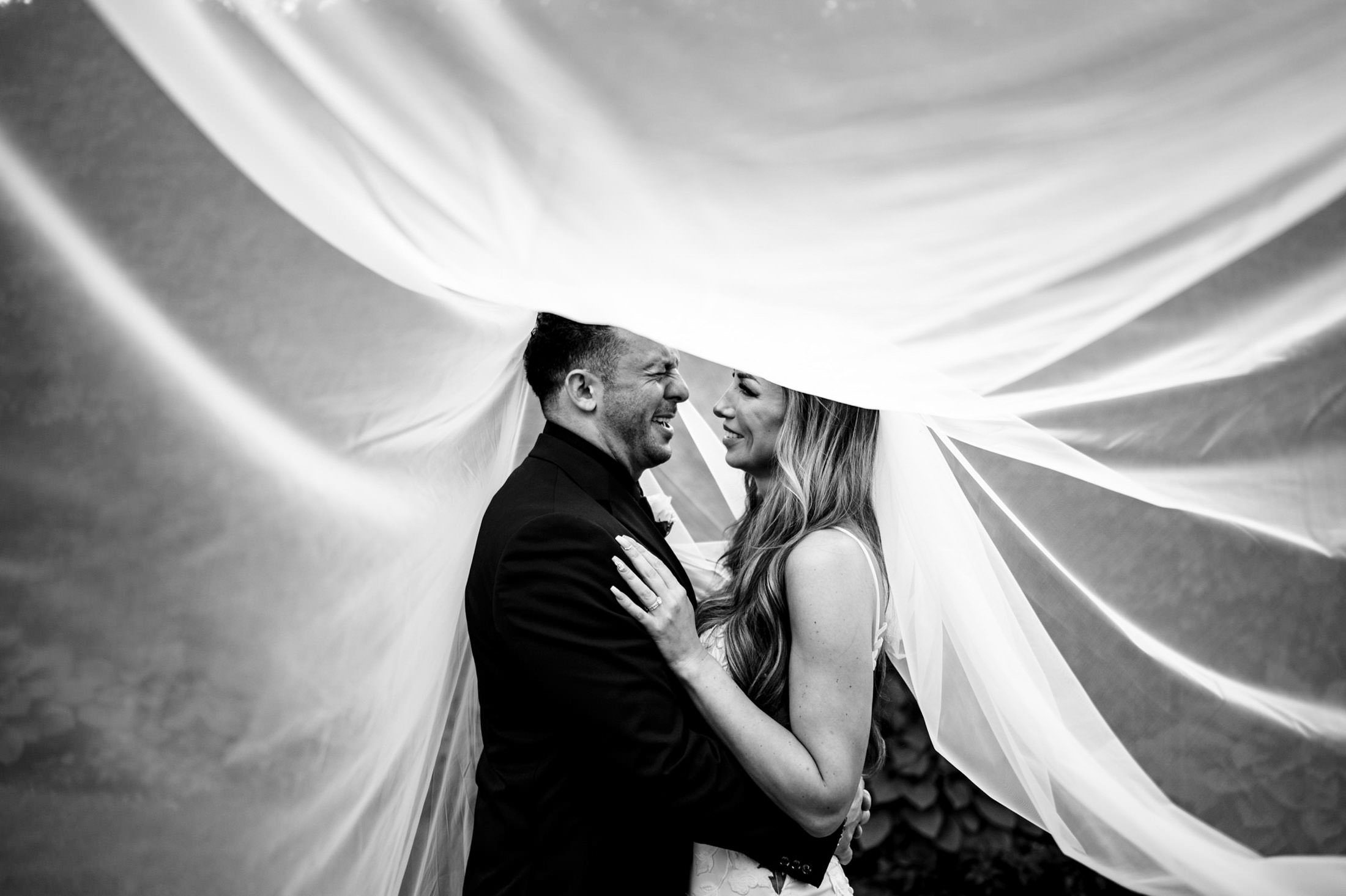 Bride and groom smiling under a flowing veil, black and white, at an Italian wedding.