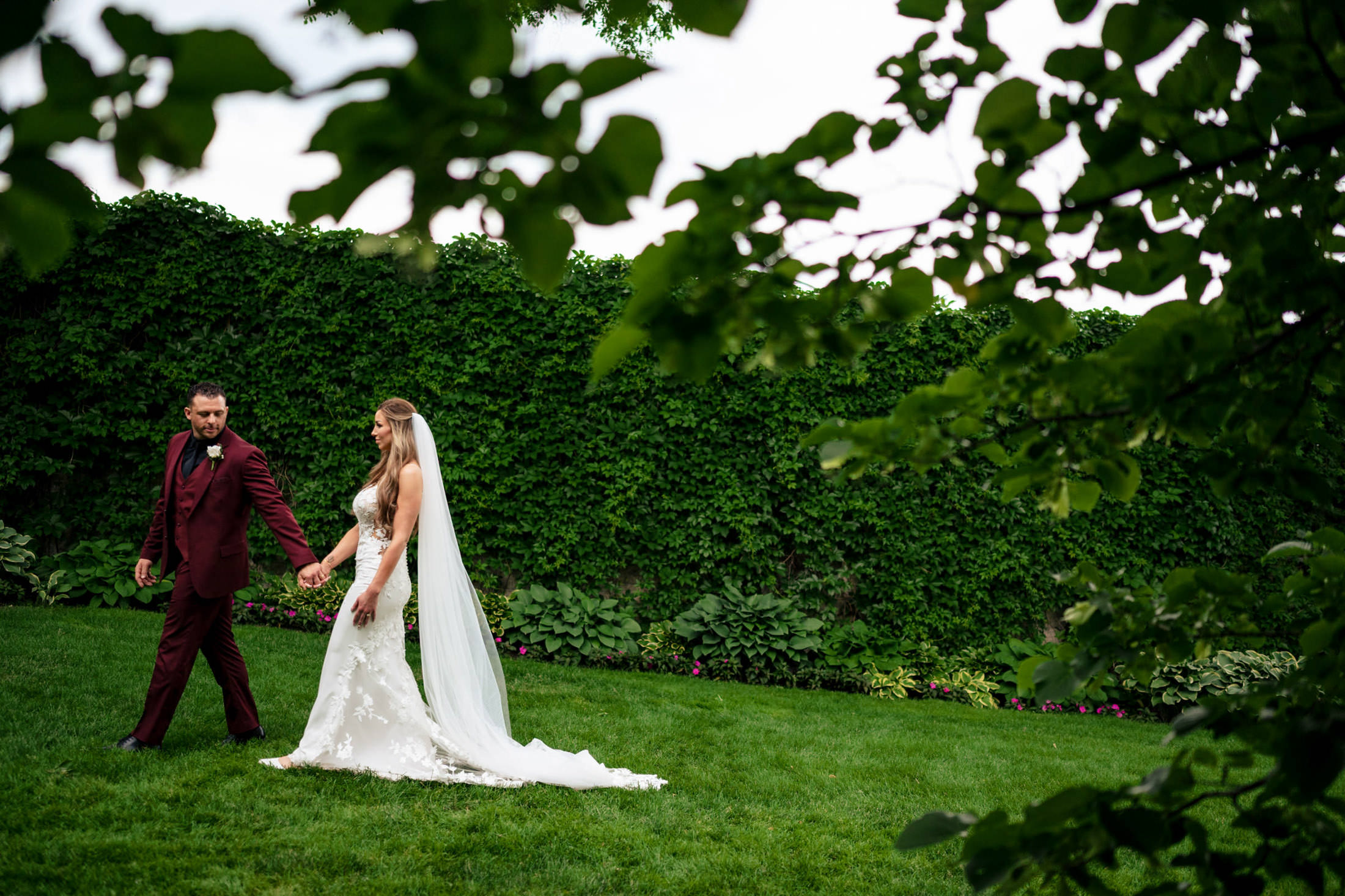 Bride and groom stroll hand in hand on grass at Italian wedding, surrounded by lush greenery.