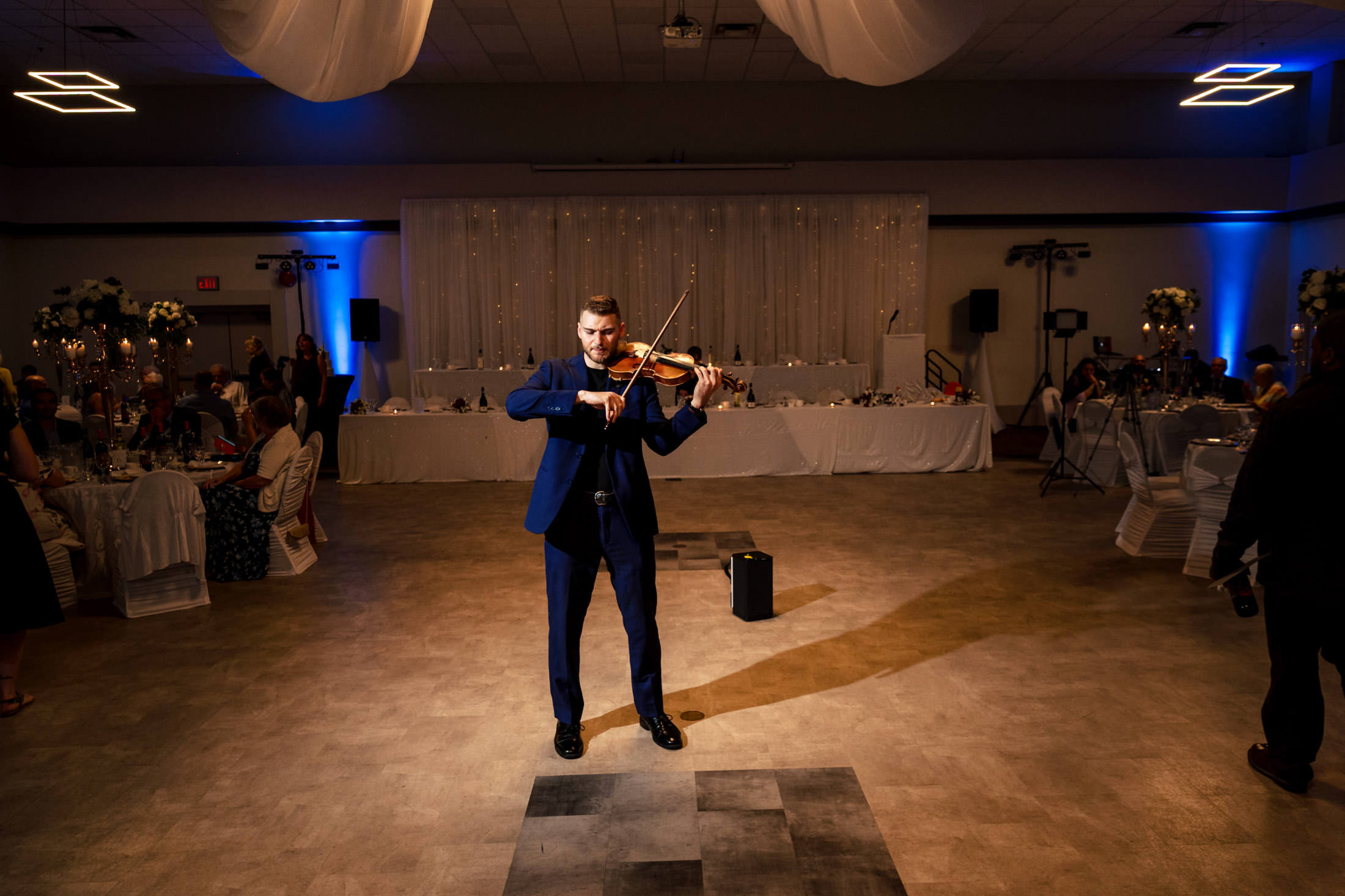 At an Italian wedding in Winnipeg, a man in a blue suit plays the violin in a dimly lit banquet hall.
