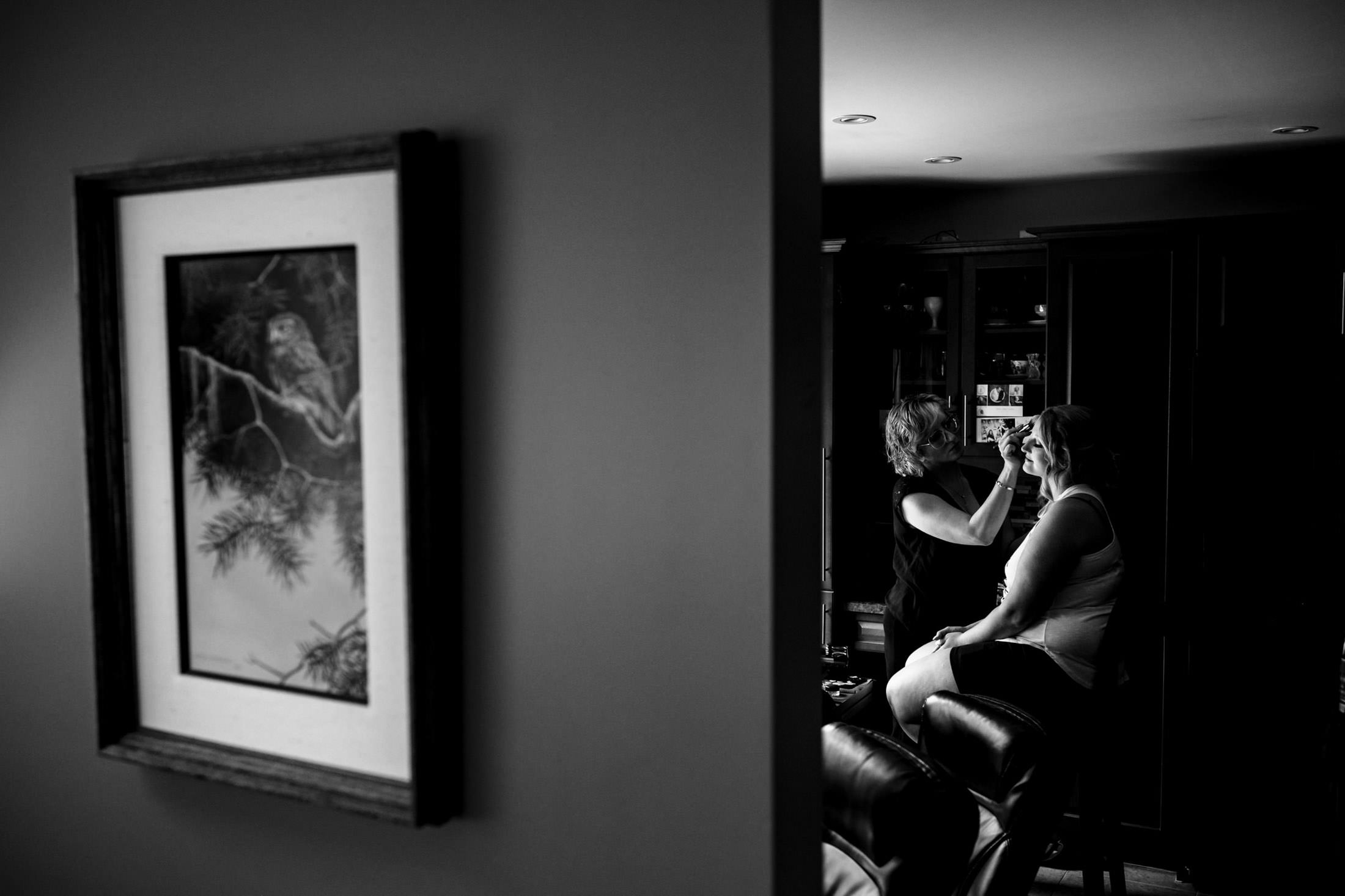 A woman applies wedding makeup in a living room; owl painting on the adjacent wall.