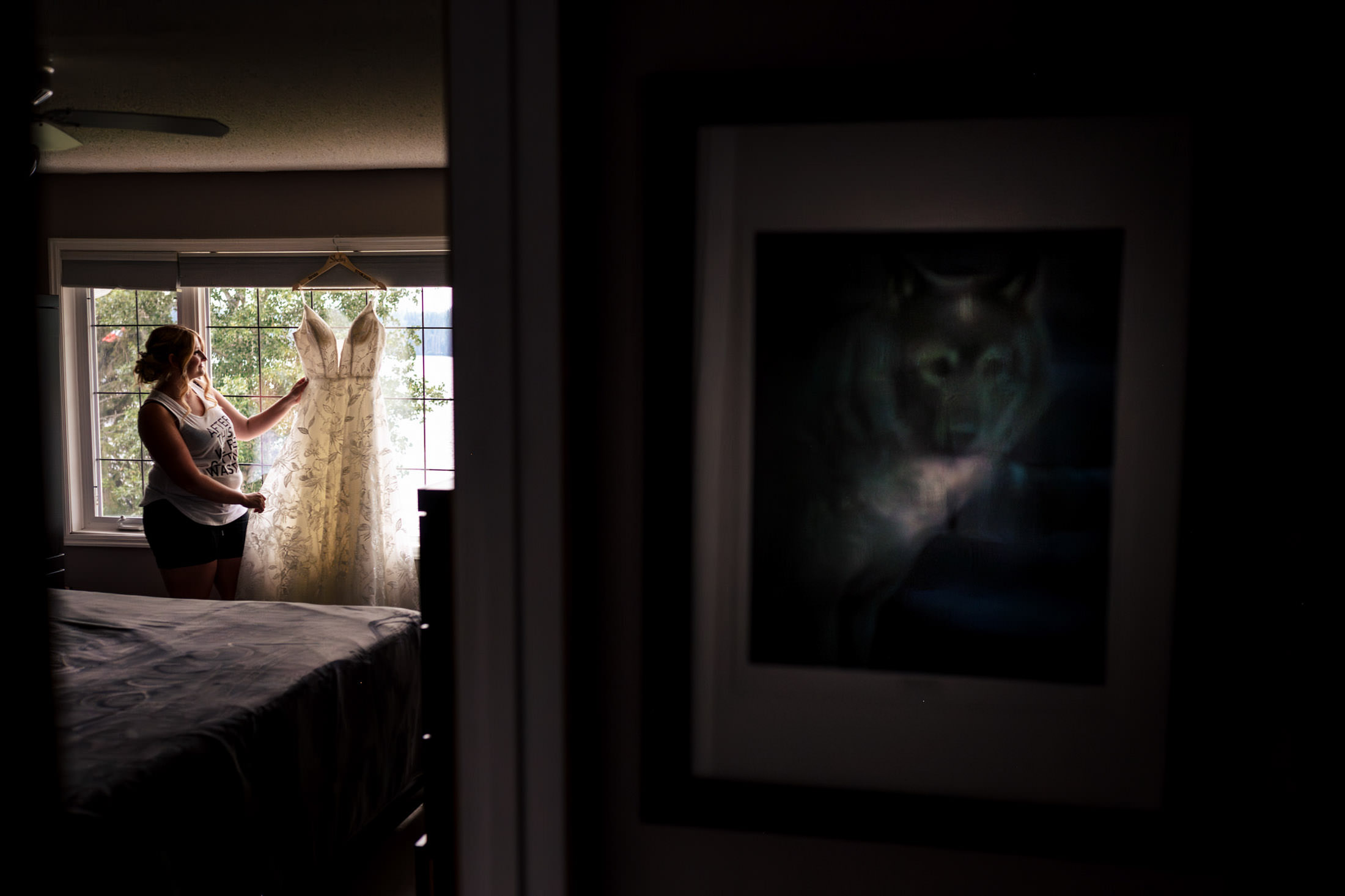 Bride admiring dress by window; wolf picture reflected on glass at Snow Lake wedding.