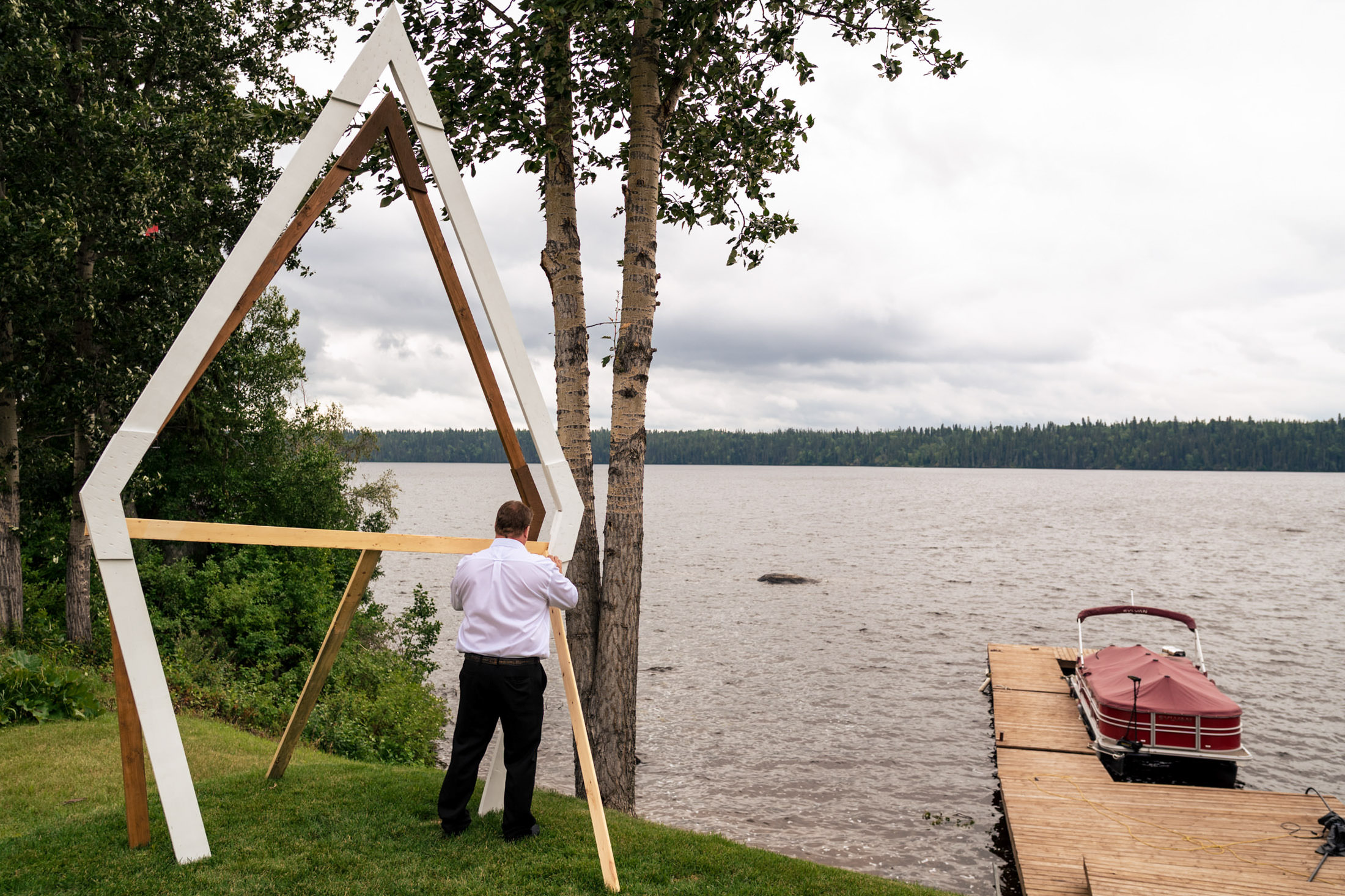 Person in white shirt near a wooden frame by Snow Lake, with a dock and boat in the background.