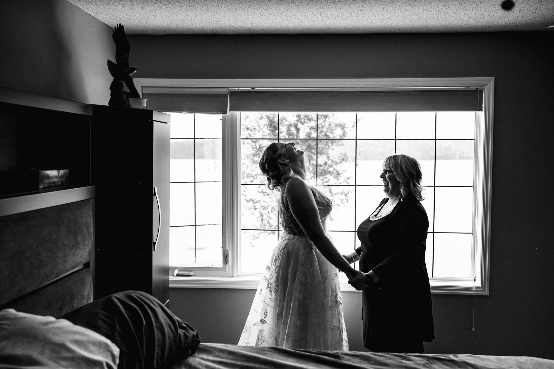 Bride and woman holding hands by a window; smiles abound at Snow Lake wedding. Black & white photo.