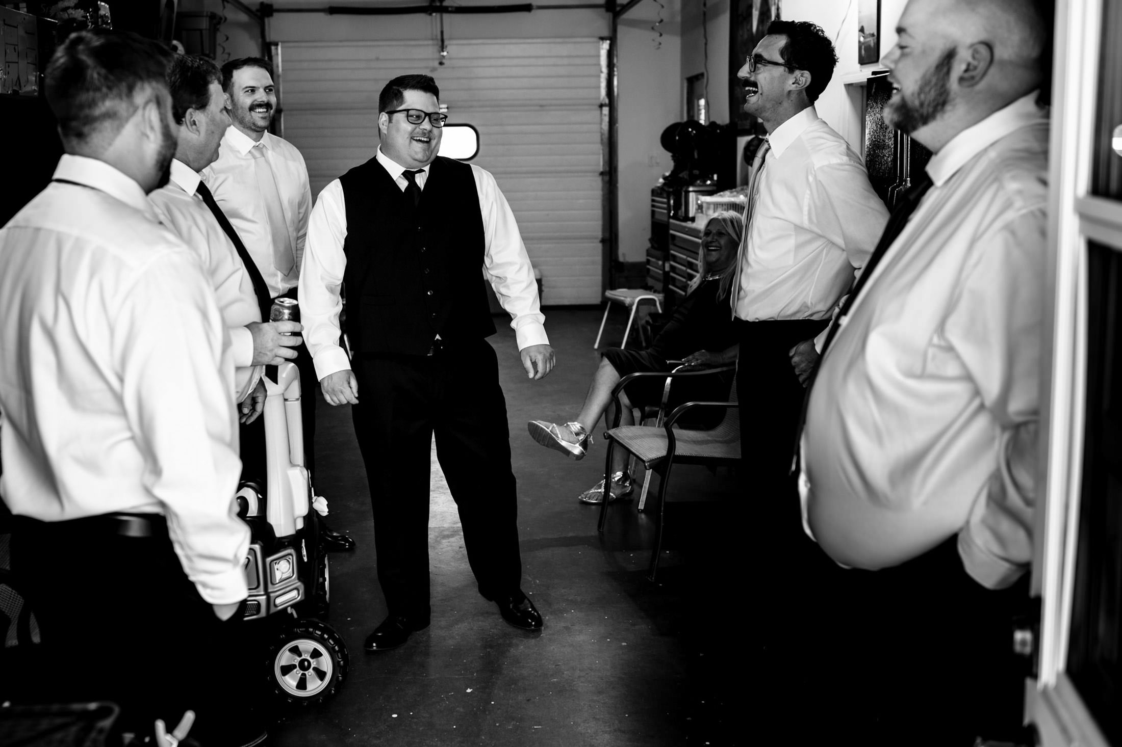 Men in formal attire share laughter in a garage, reminiscing about a wedding at Snow Lake.