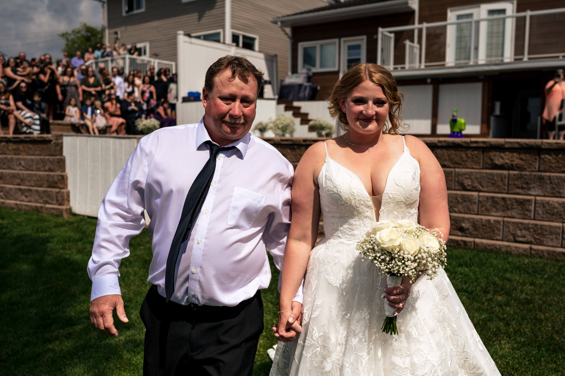 Bride walking with a man in a white shirt at Snow Lake, holding a bouquet, guests in the background.