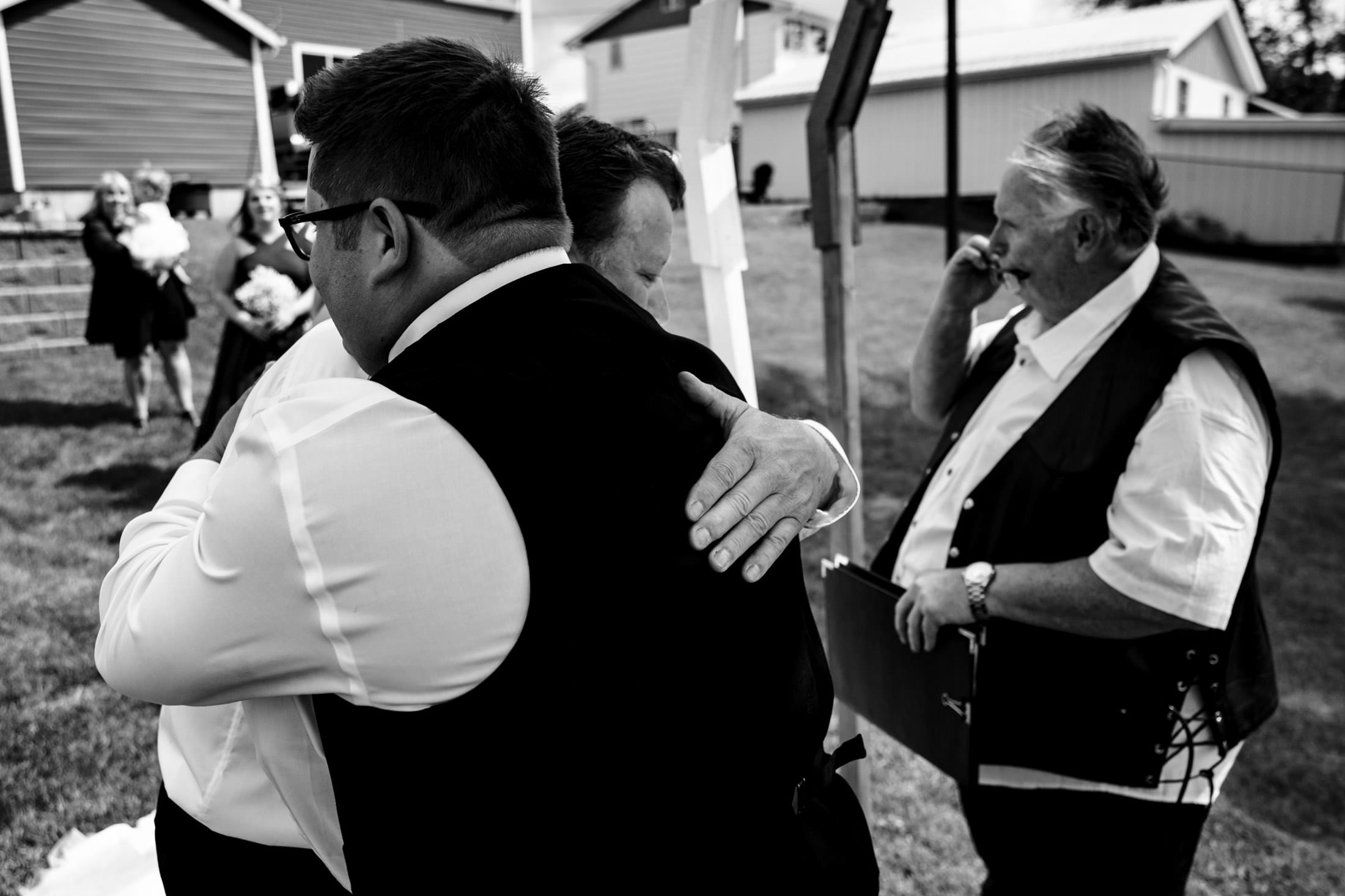 Two people hugging at Snow Lake wedding; a man in a vest talks on the phone nearby.