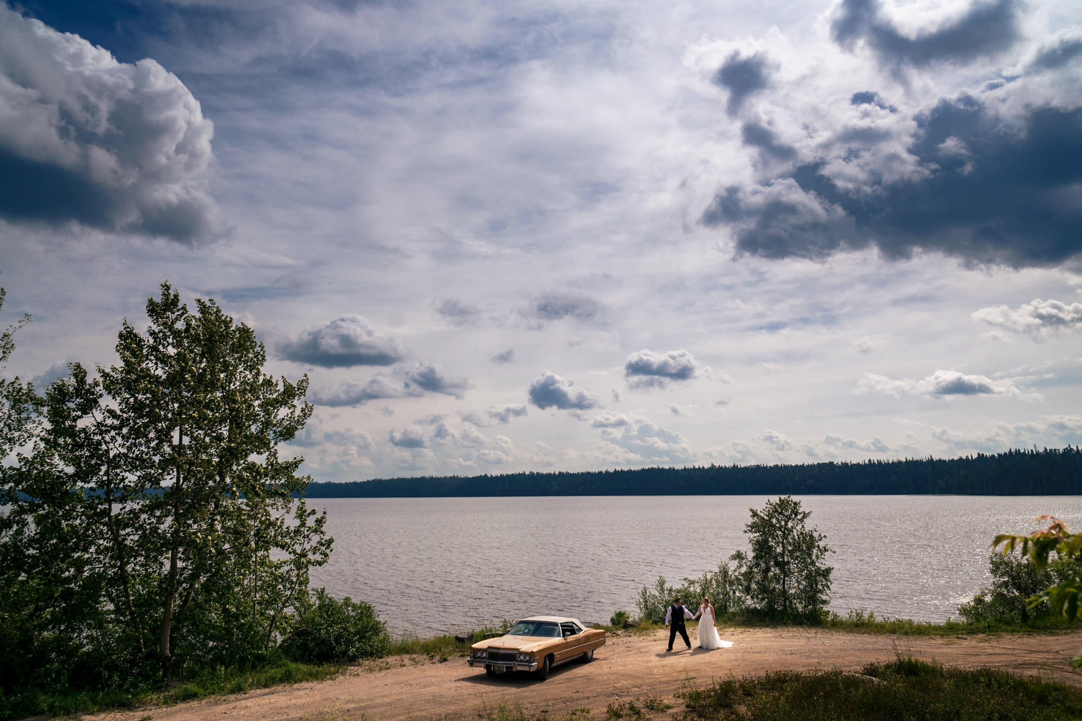 Bride and groom by Snow Lake with a vintage car under a cloudy sky.