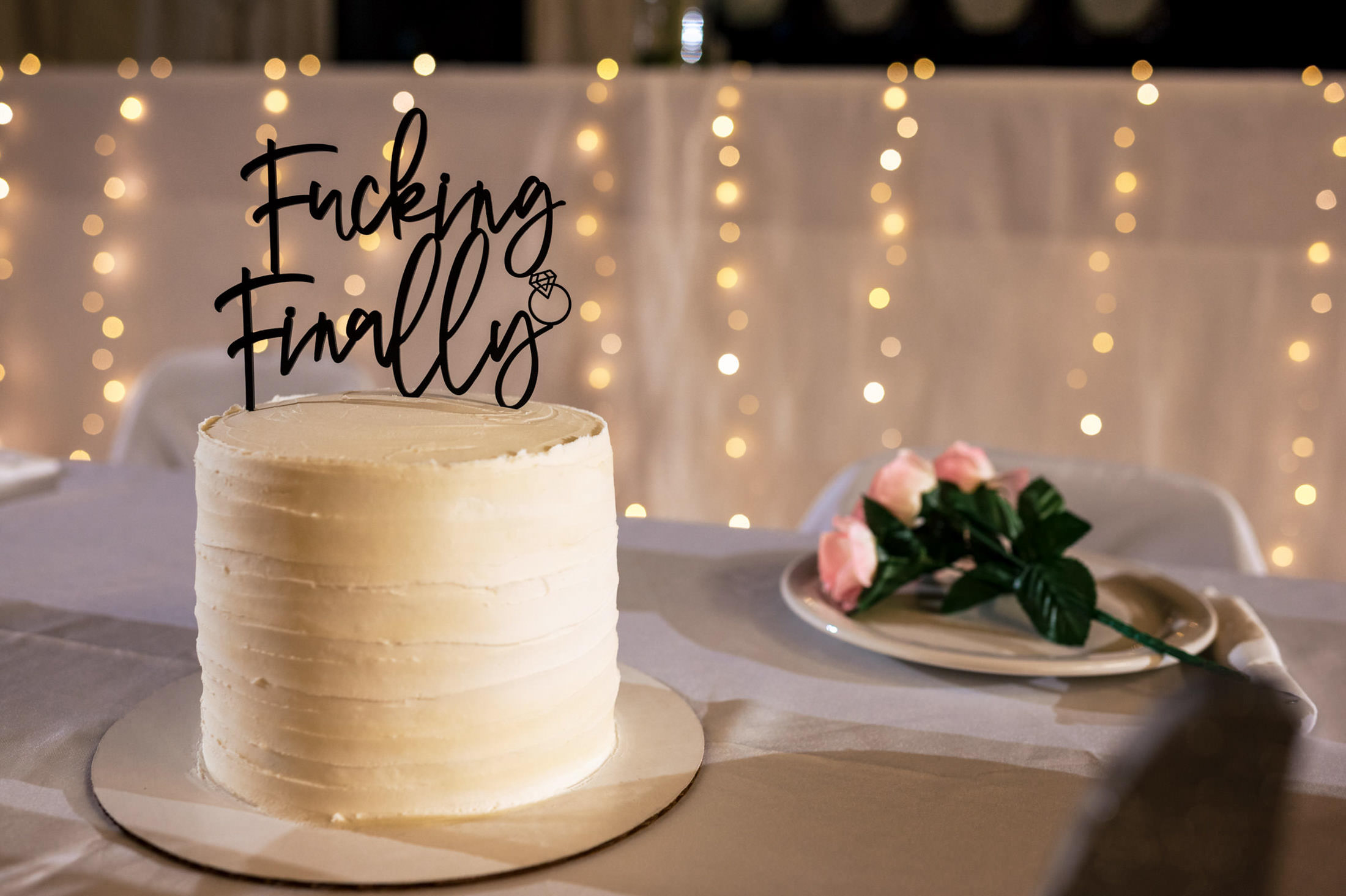 White cake with a "Fucking Finally" topper and pink roses, perfect for a wedding at Snow Lake.