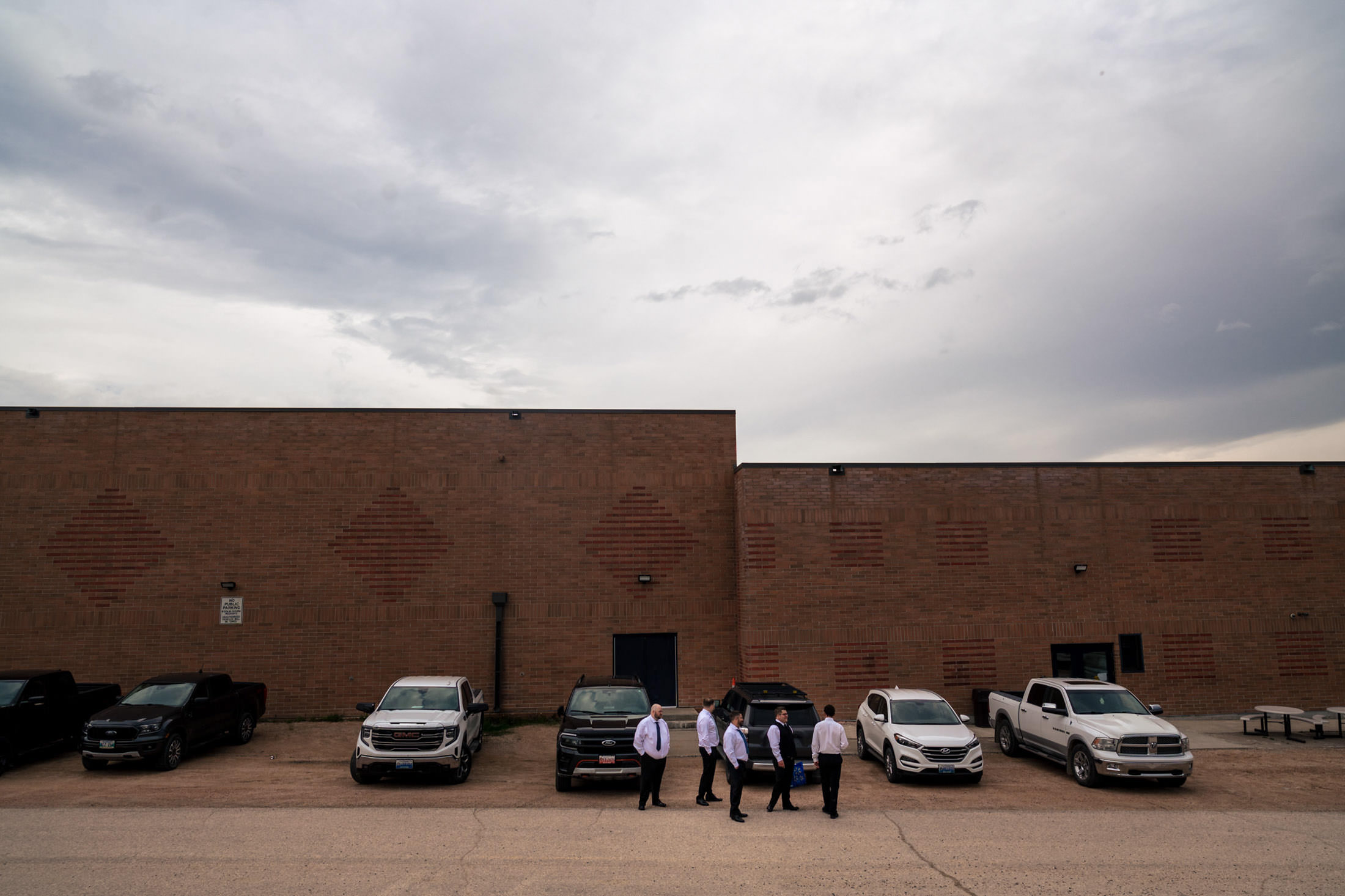 Five people in white shirts stand by trucks outside a brick building near Snow Lake under clouds.