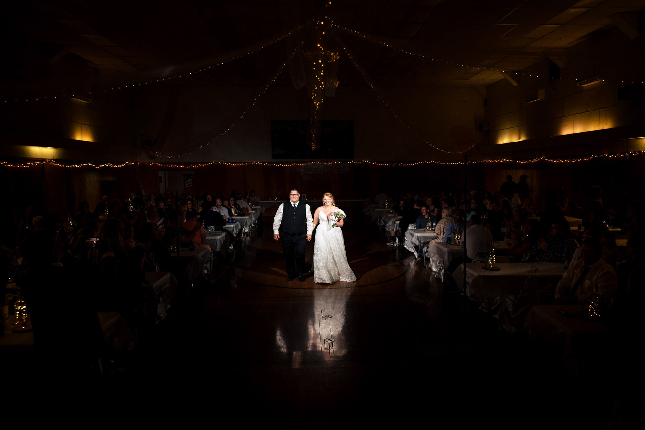 Bride and groom stroll at Snow Lake venue, guests seated in a dimly lit ambiance.