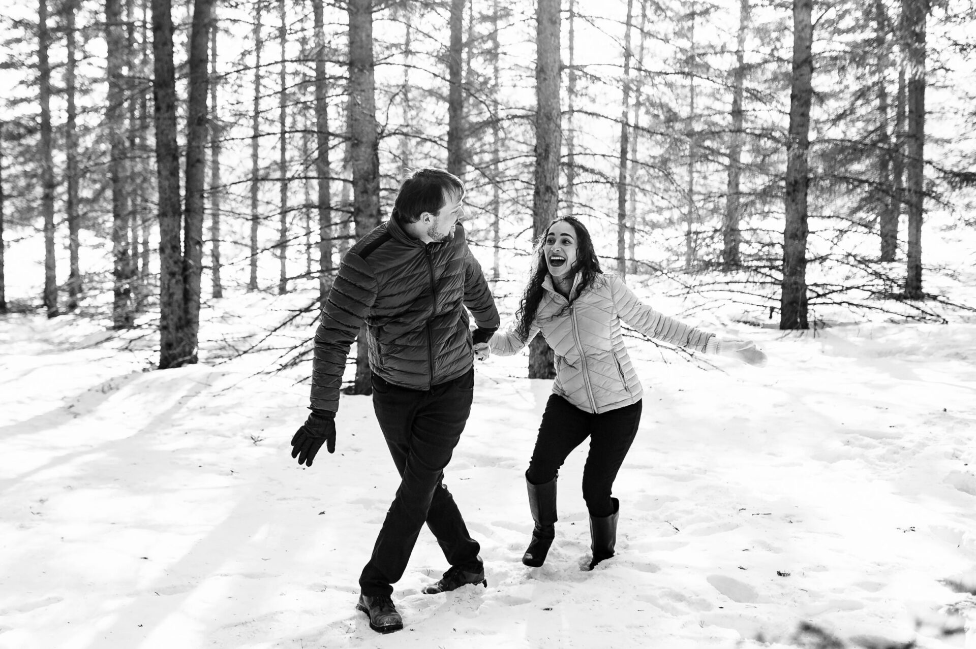 A joyful couple in Winnipeg's snowy forest, smiling and holding hands.