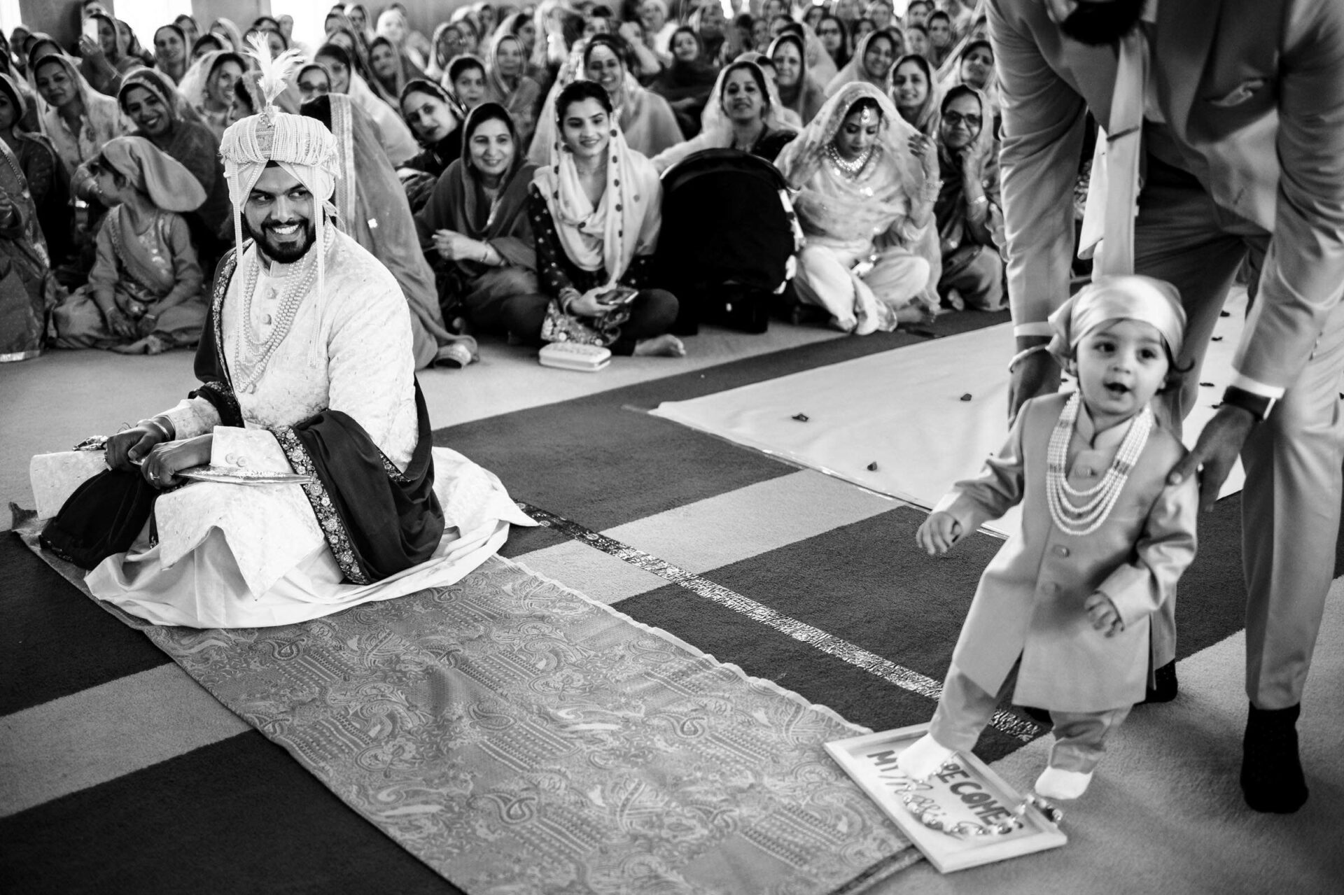 A groom smiles as a child in traditional attire walks forward—a best Winnipeg wedding photo.
