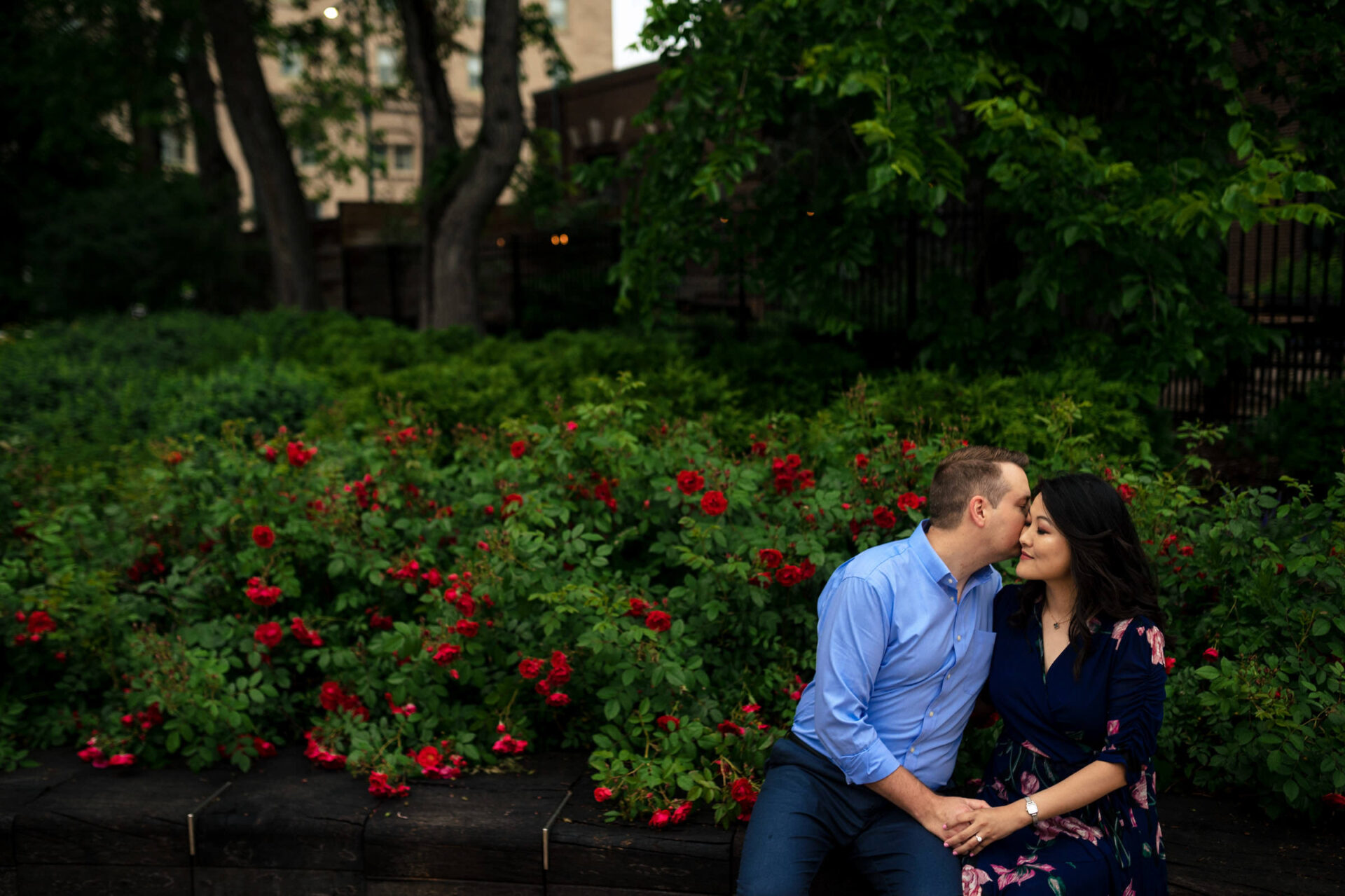 A couple shares a tender moment on a bench, lush greenery, and red blooms frame their joy.