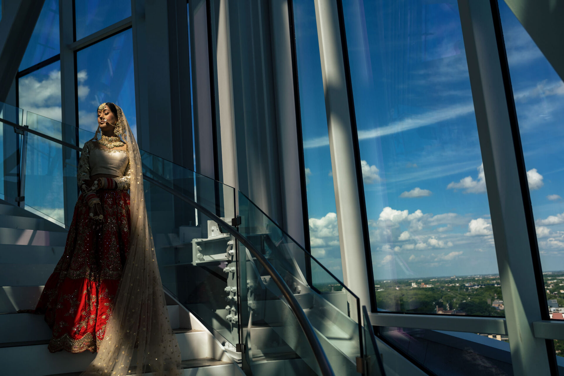 Bride in traditional attire descends a glass staircase with Winnipeg's skyline in view.