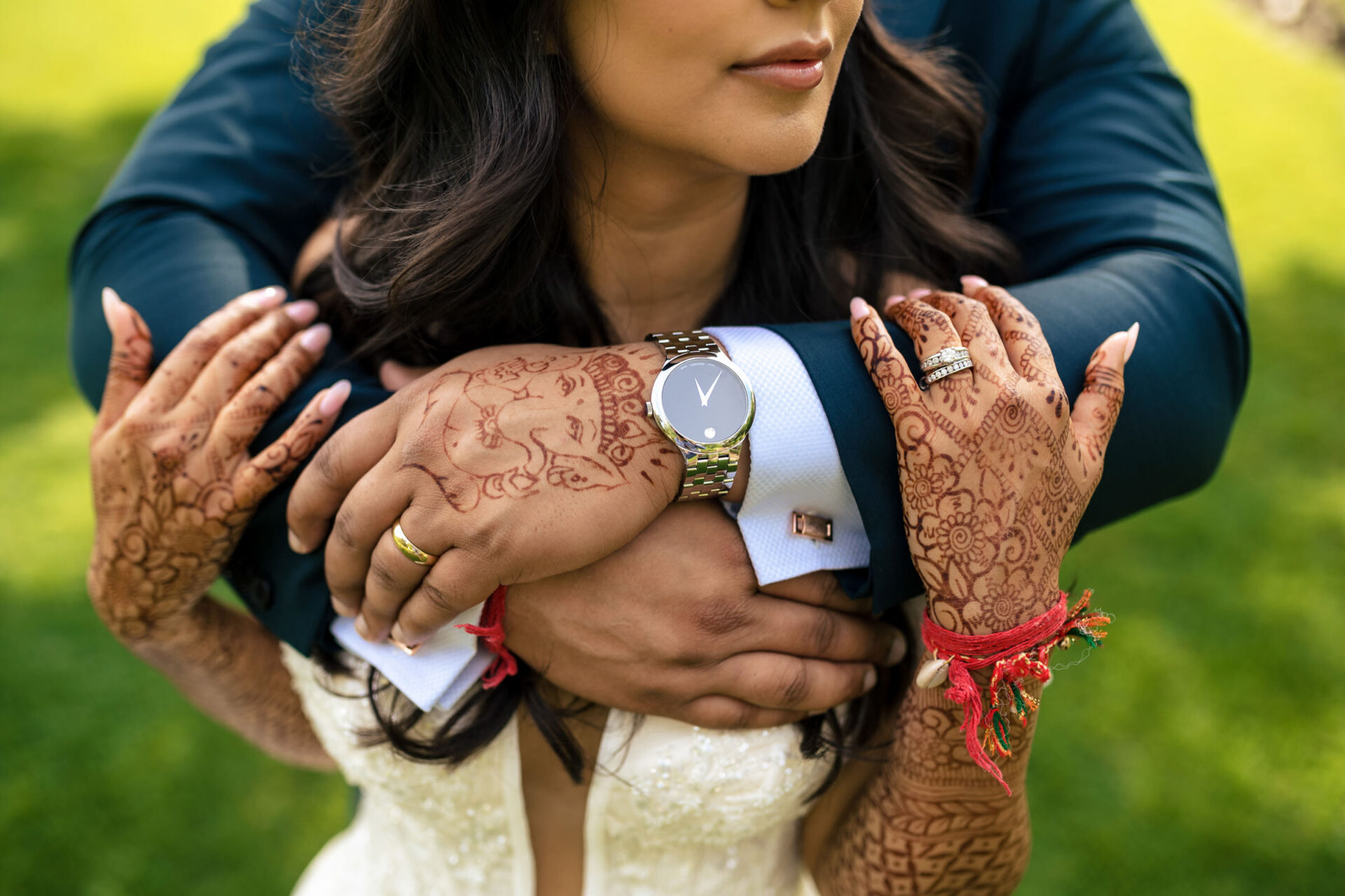 Bride's henna-touched hands hold groom's, with rings and watch, over lush green grass.