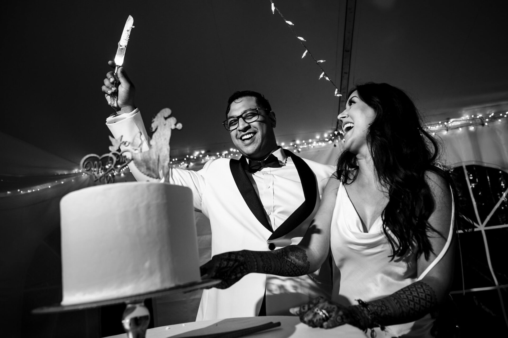 Best Winnipeg Wedding Photos: Couple cutting cake under string lights, smiling brightly.