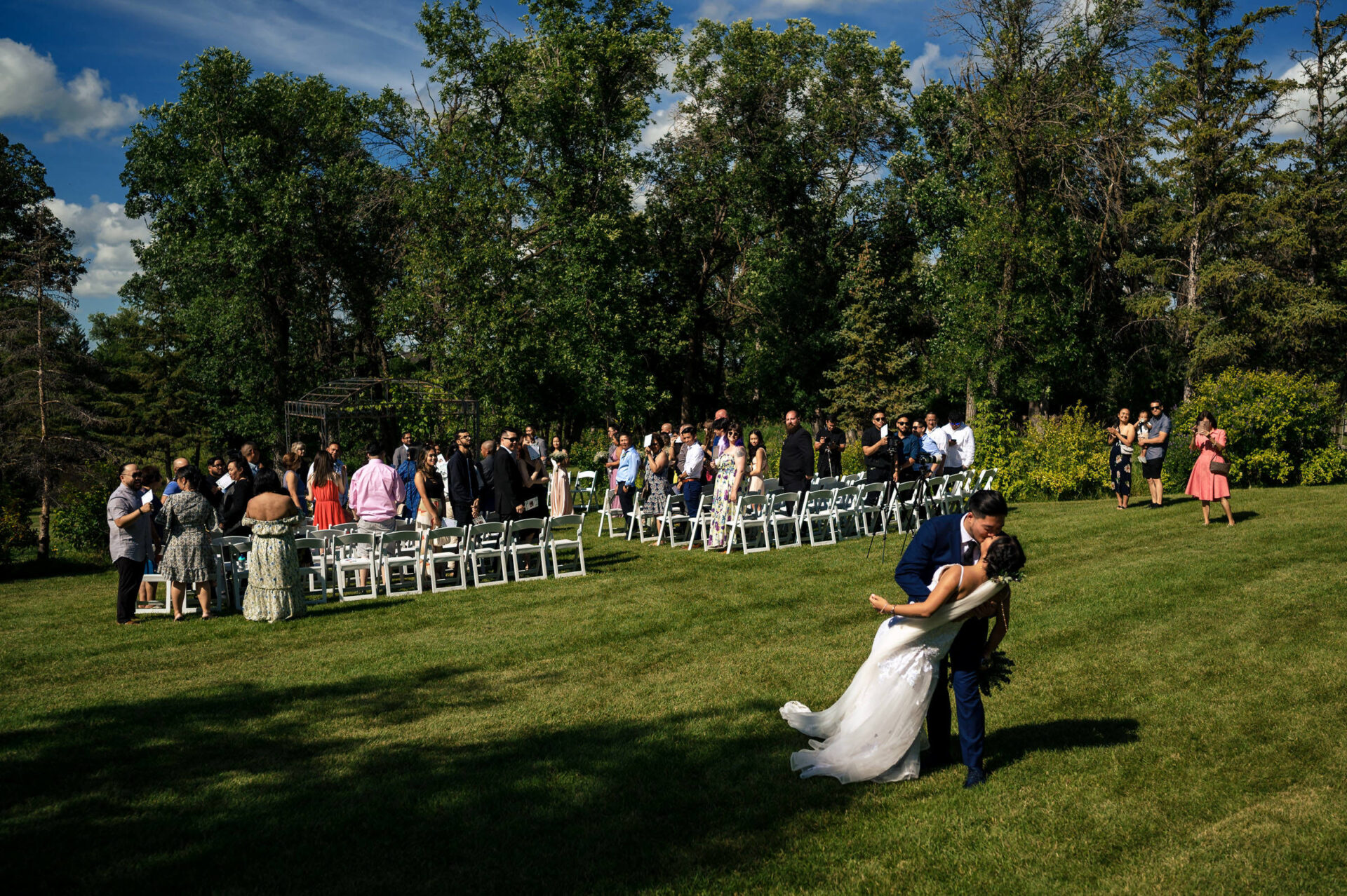 A couple kisses on the grass as guests gather in a wooded setting for best Winnipeg wedding photos.