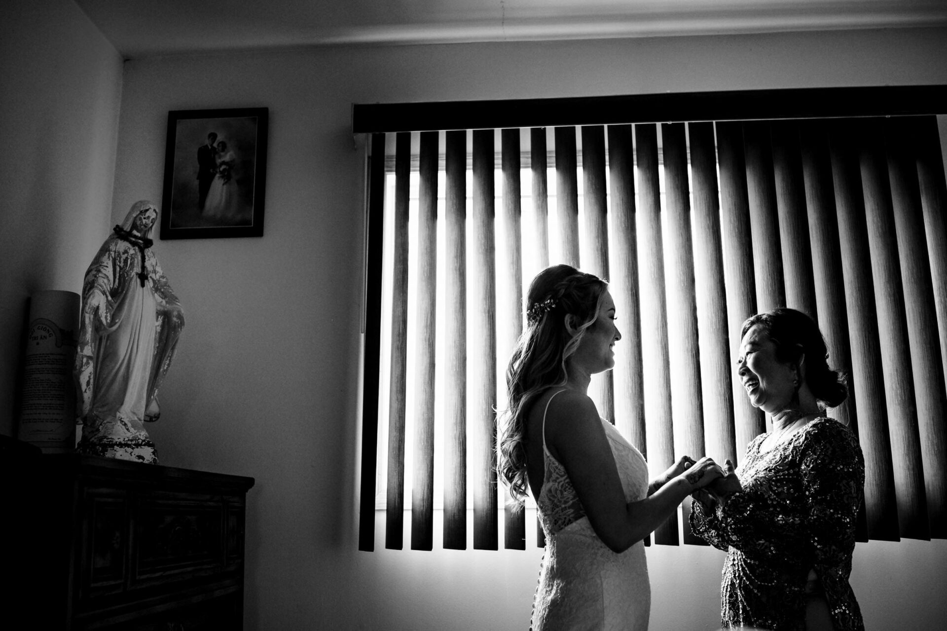 Bride holds hands with woman by window blinds, a perfect moment in the best Winnipeg wedding photos.