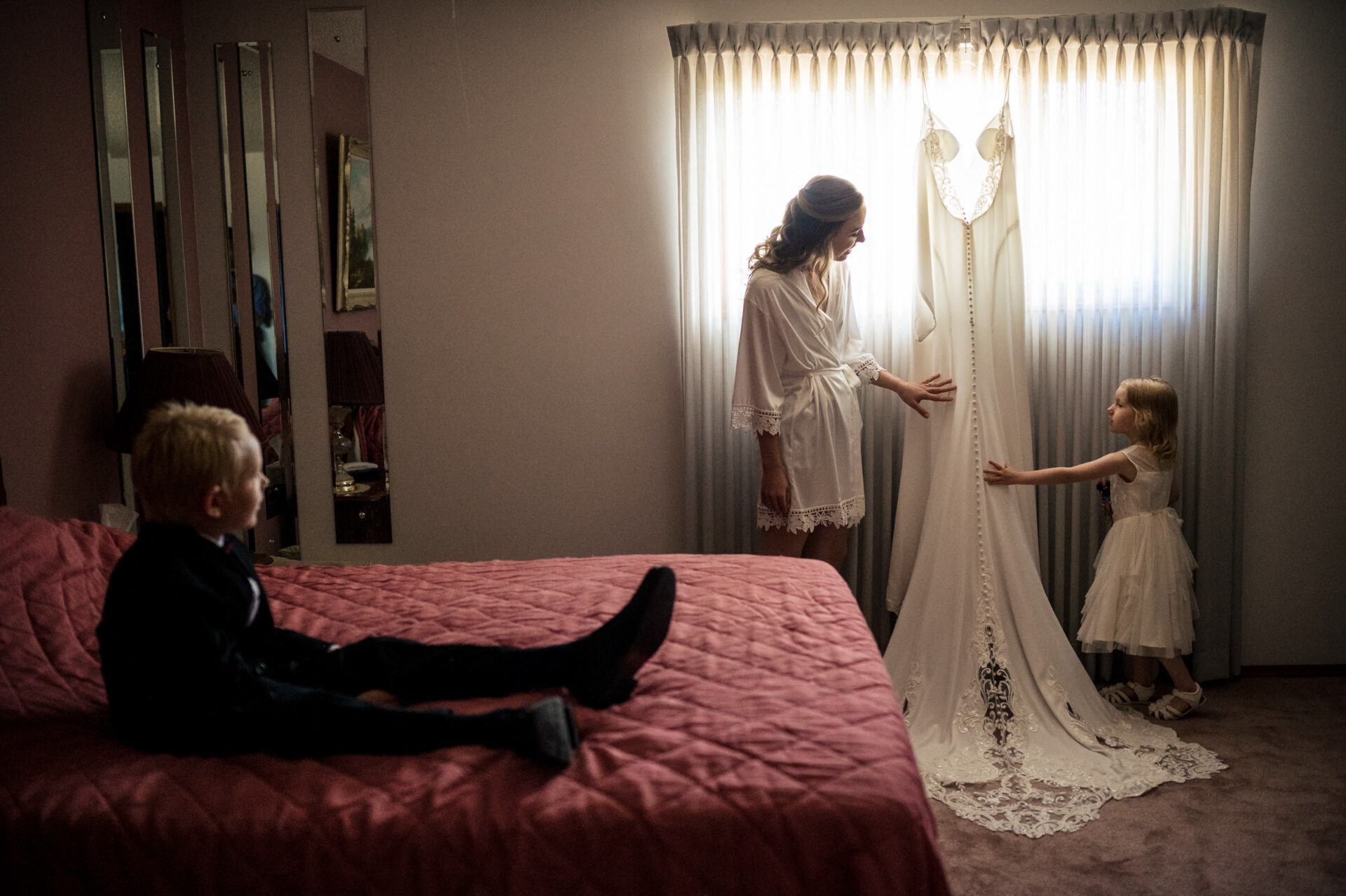 Bride and child admire dress; boy sits on bed, a perfect Winnipeg wedding photo moment.