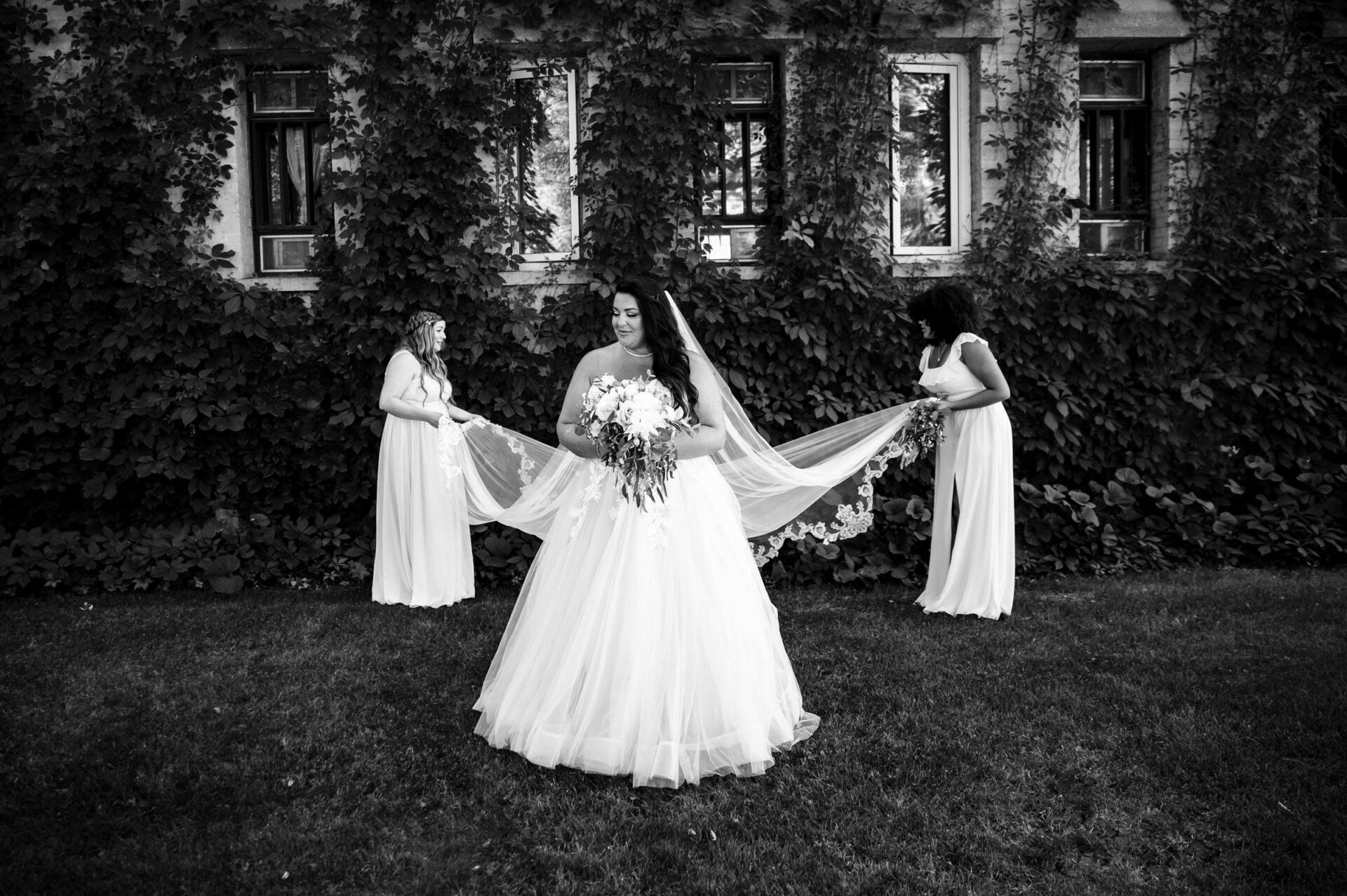 Bride in gown outdoors, two bridesmaids adjust her veil, best Winnipeg wedding photo.