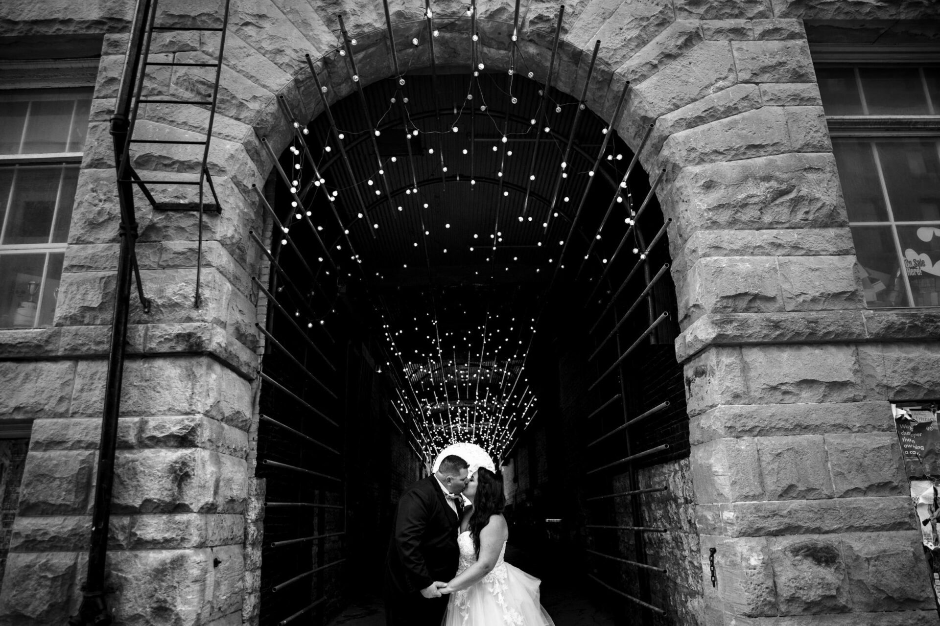 Bride and groom kiss under twinkling lights in Winnipeg's stone archway.