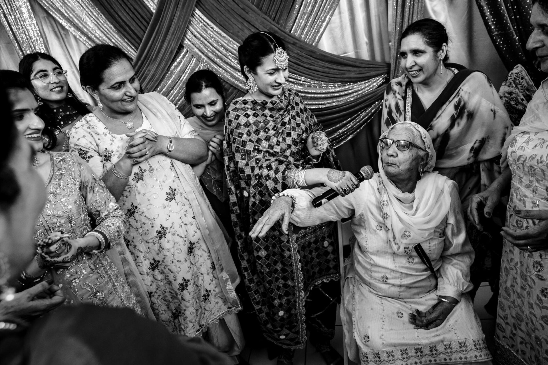 Elderly woman speaks into microphone, surrounded by smiling women in traditional Winnipeg attire.