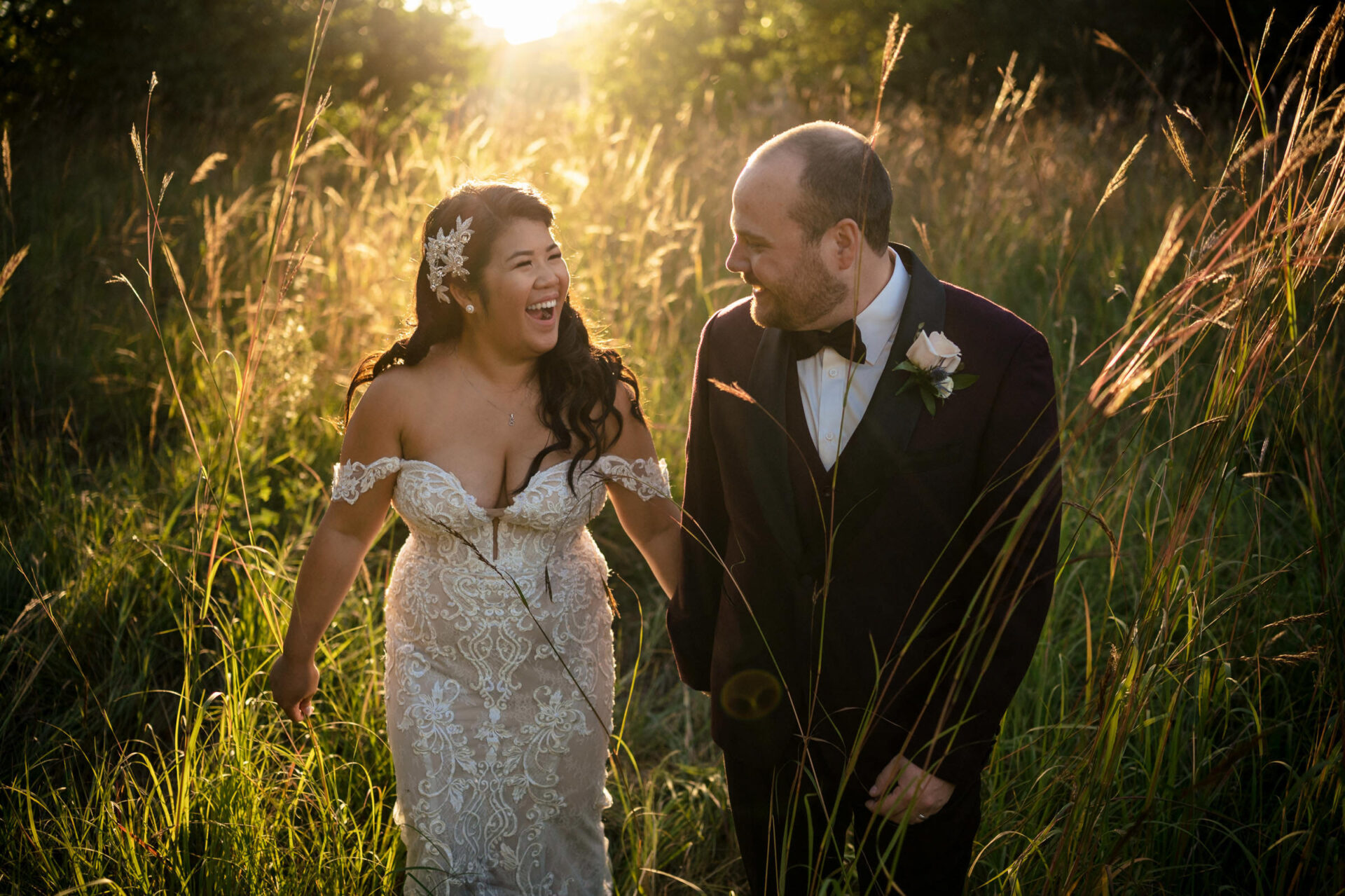 Bride and groom smiling, walking through tall grass in Winnipeg sunlight.