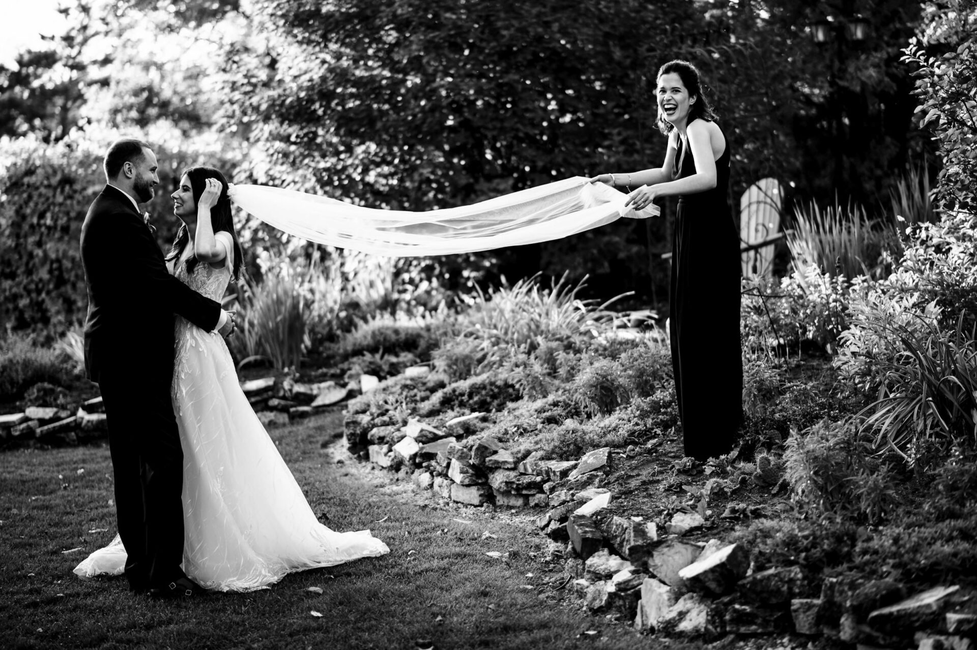 Bride and groom smile as friend holds veil; garden setting, Winnipeg wedding photo.
