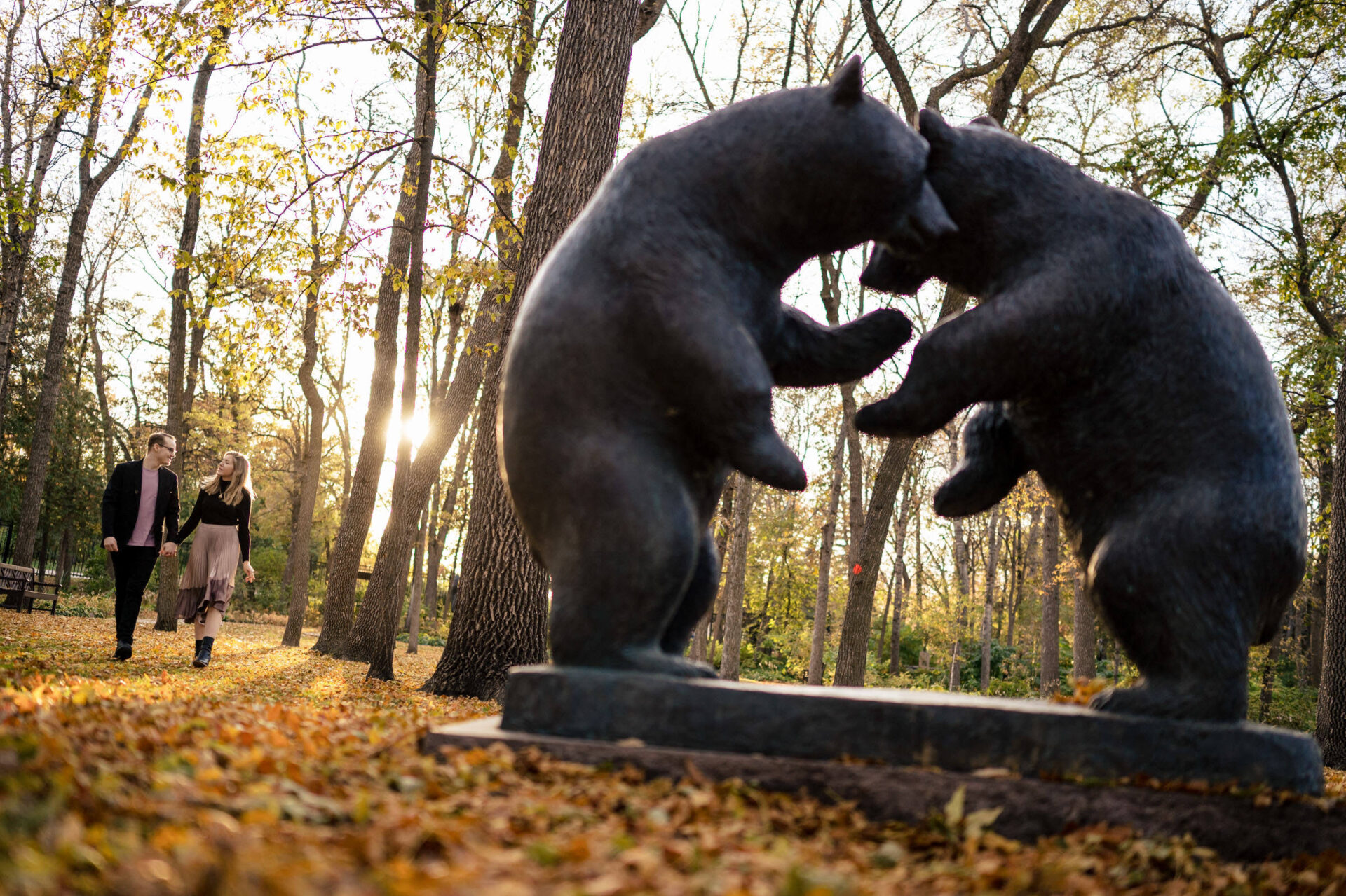 Two people walk near a statue of bears in a sunlit, autumn forest—best Winnipeg memories.