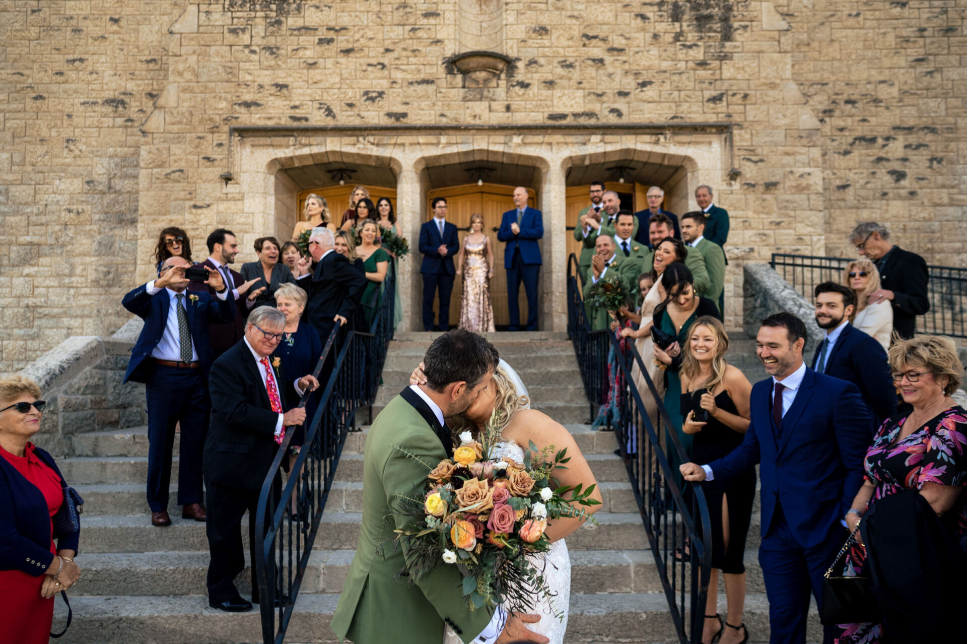 A couple kisses on stone steps, in one of the best Winnipeg wedding photos, cheered by loved ones.
