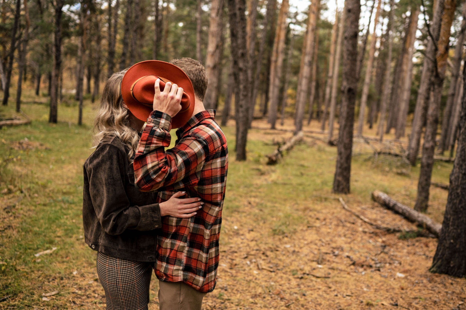 Couple in woods kisses, man holds hat, hiding their faces; best Winnipeg wedding photo vibe.