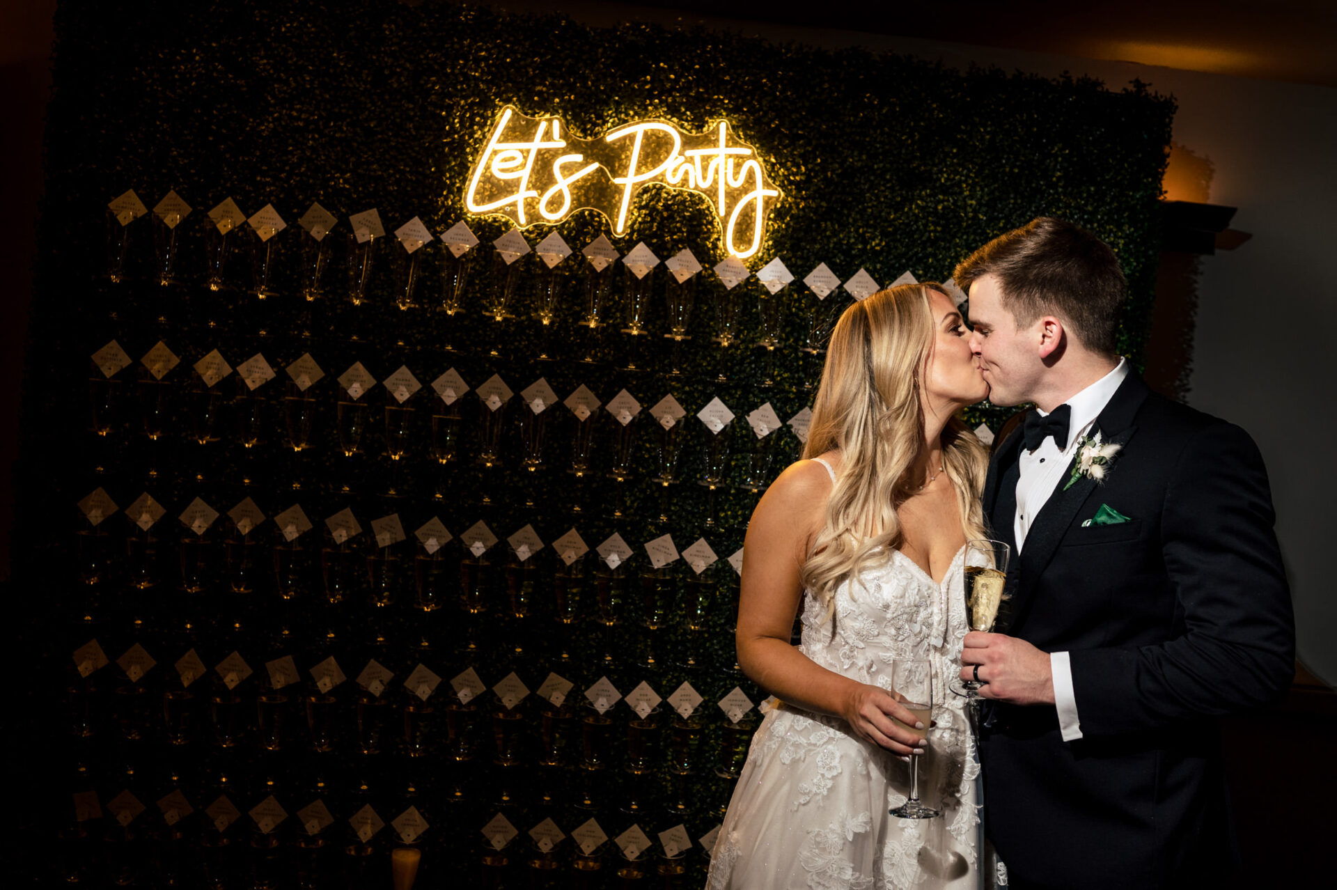 A bride and groom kiss, with champagne, under a "Let's Party" neon in the Best Winnipeg Wedding Photos.