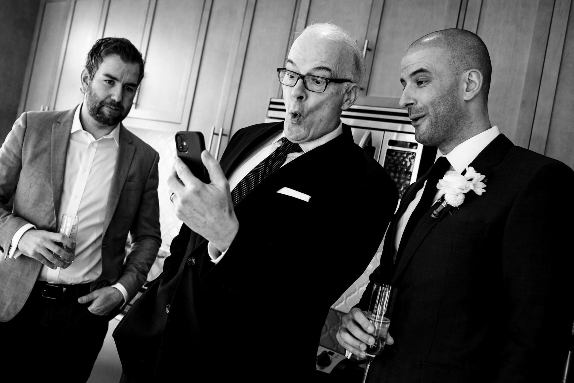 Three men in suits react to a phone, two with champagne, capturing Winnipeg's best wedding moments.