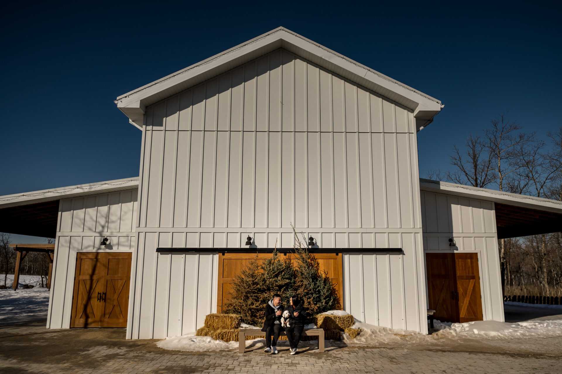 Two people sit on a bench by a large white barn, celebrating their winter engagement.