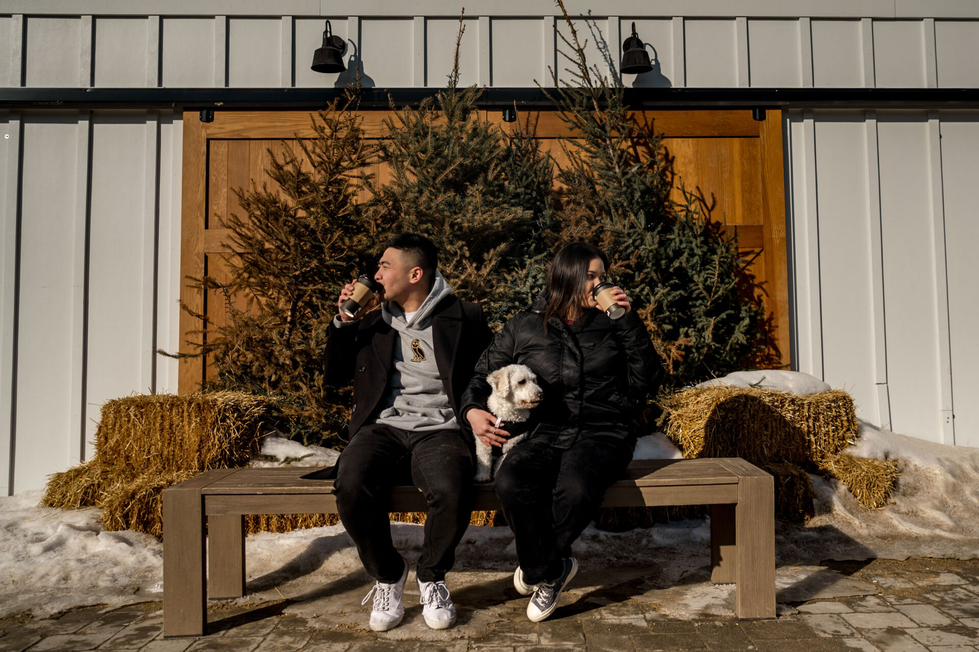 Two people on a bench, sipping coffee with their dog, amidst hay bales and winter's charm.