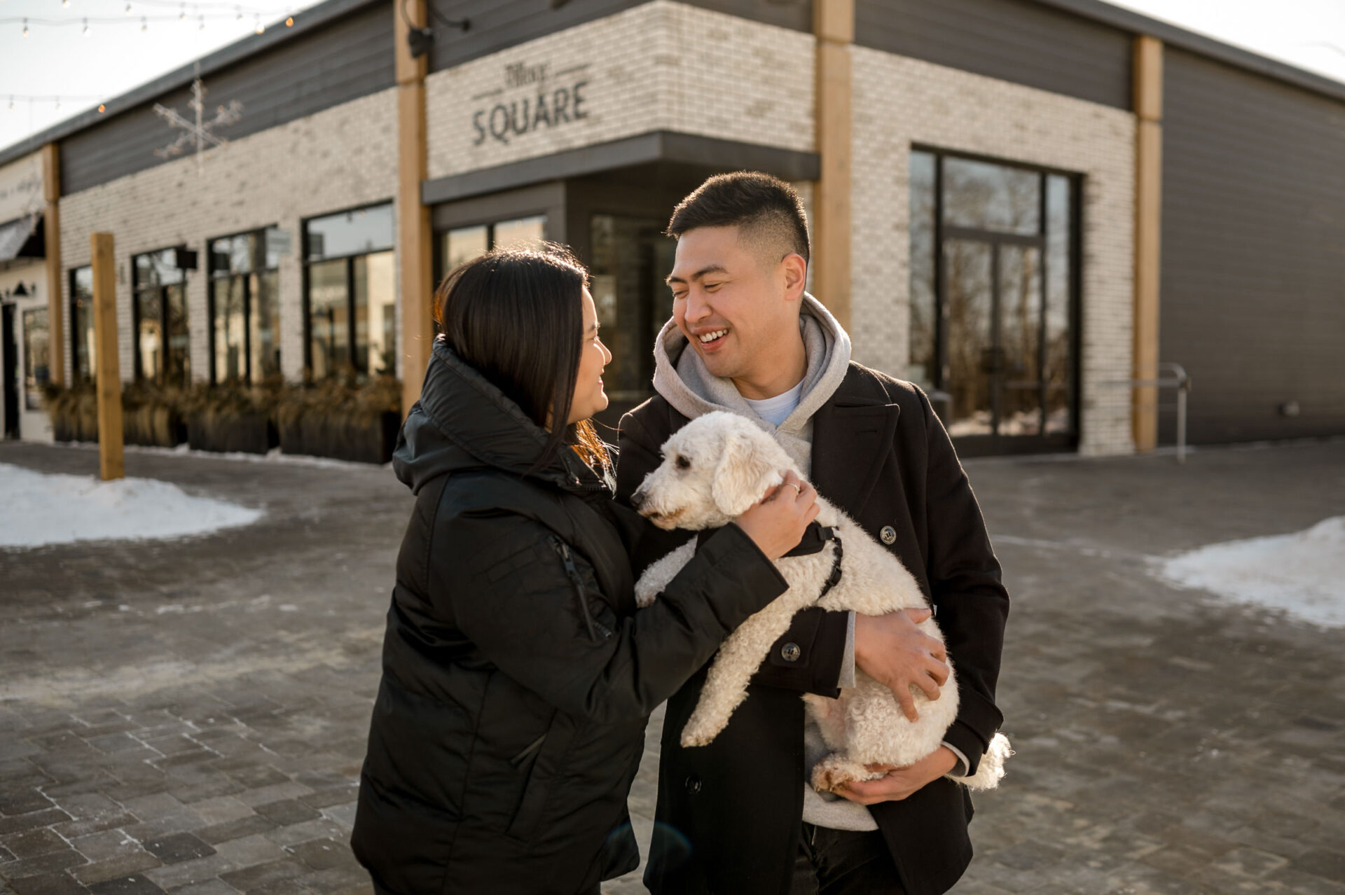 A couple shares a winter engagement moment, smiling with their small dog in the snow.