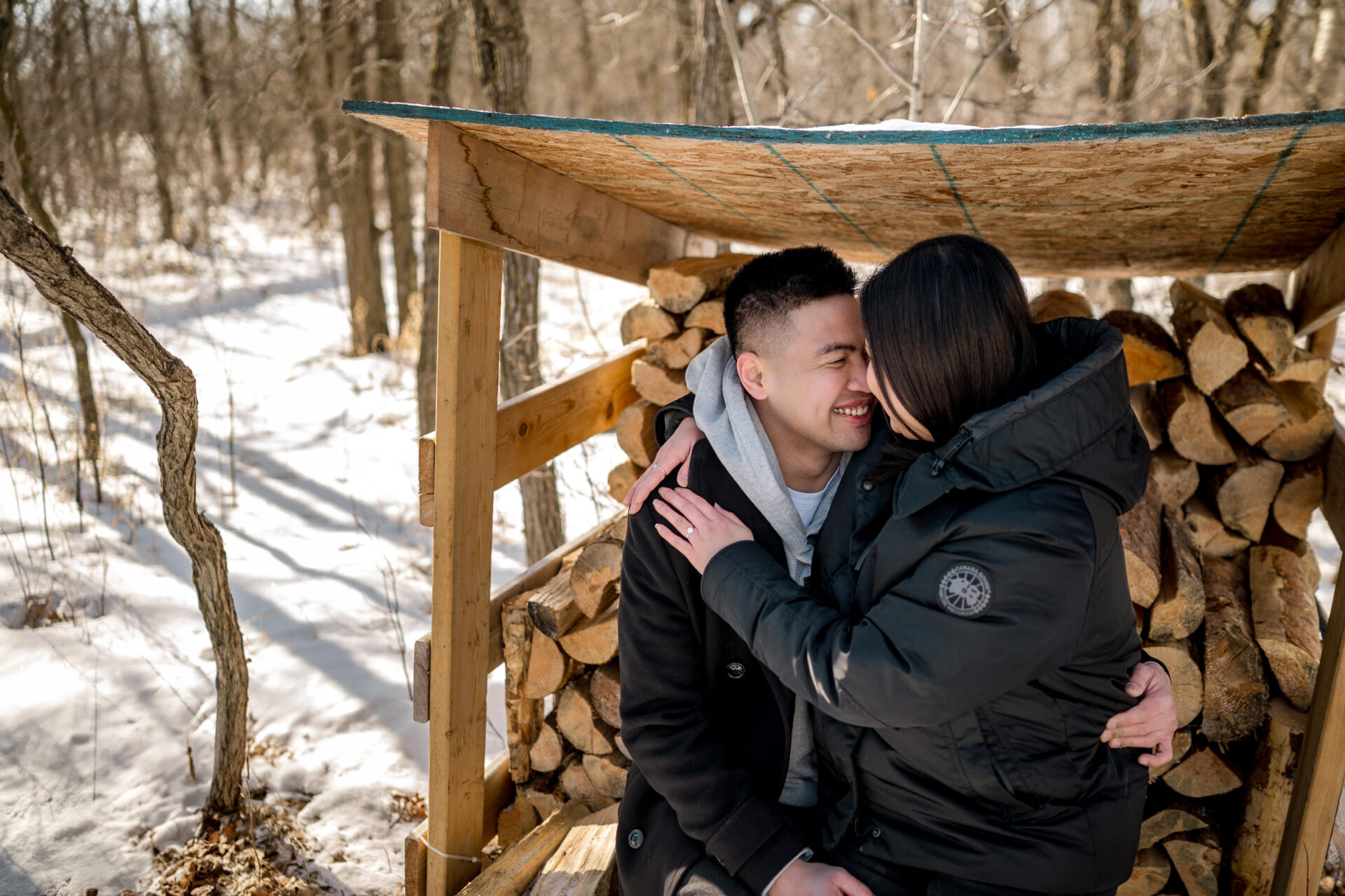 Couple embracing on a wooden bench in snowy woods, celebrating their winter engagement.