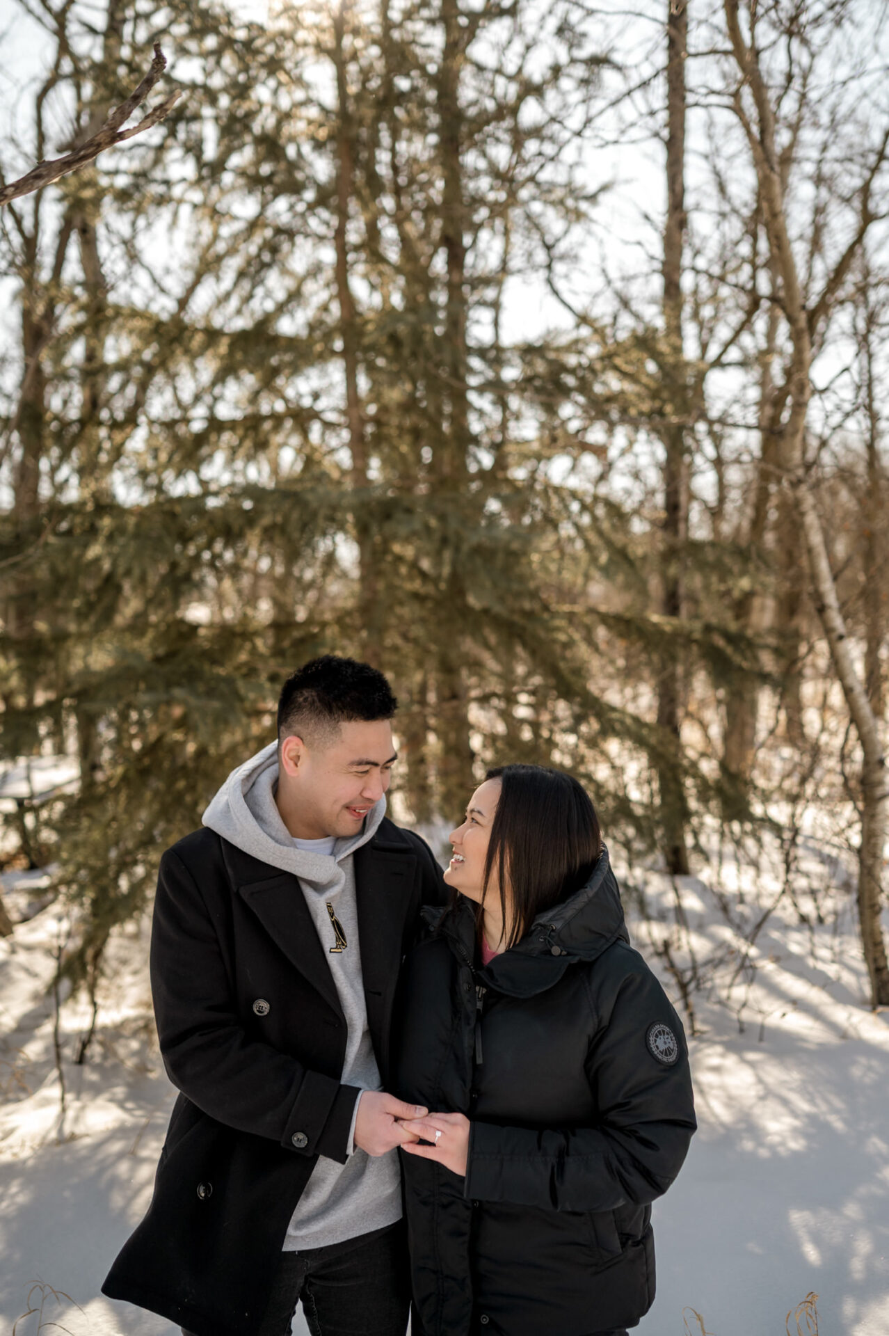 Couple smiling in snowy forest, wearing winter jackets, sharing a warm winter engagement moment.